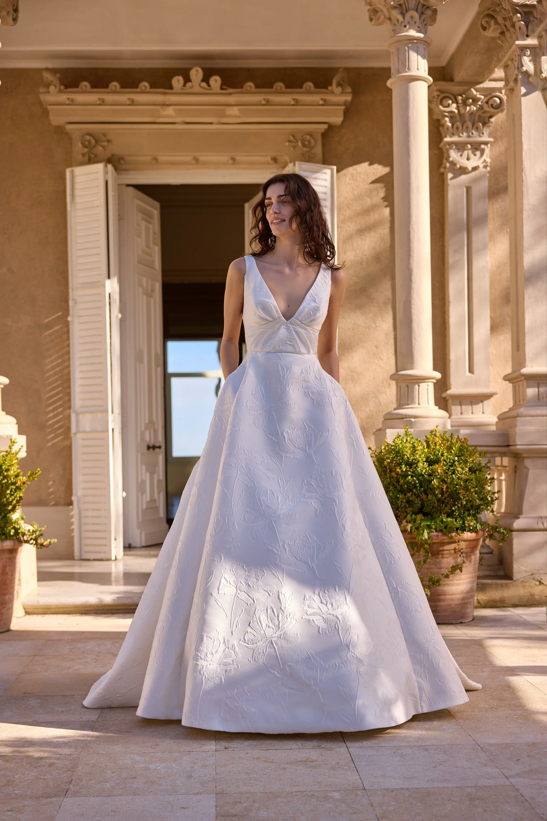 Jesus Peiro at New York Bridal Market. A woman in a white wedding dress standing outdoors in front of a building with ornate columns and open doors, bathed in warm sunlight.