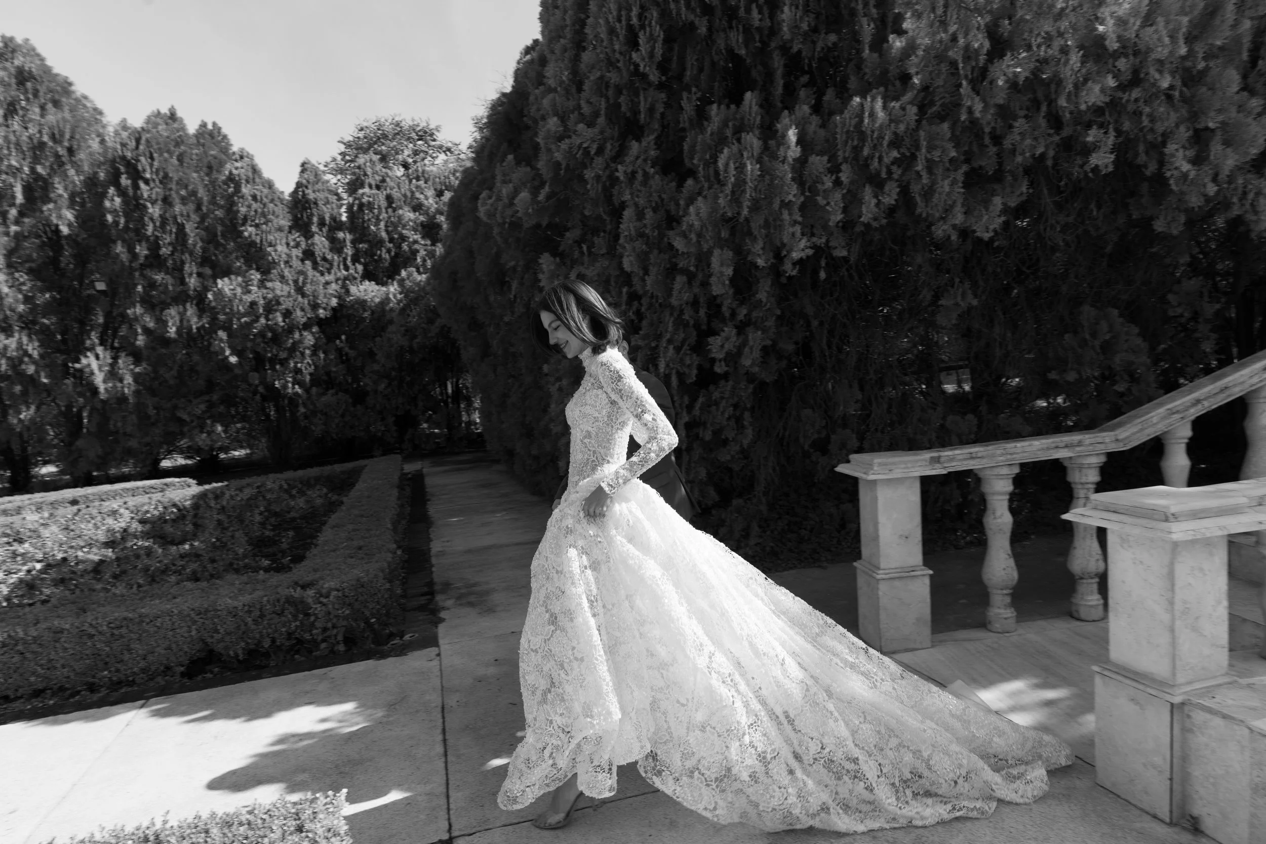 	
Guardiola at New York Bridal Market A woman in a wedding dress walking outdoors, smiling, with large trees and a stone balustrade in the background.