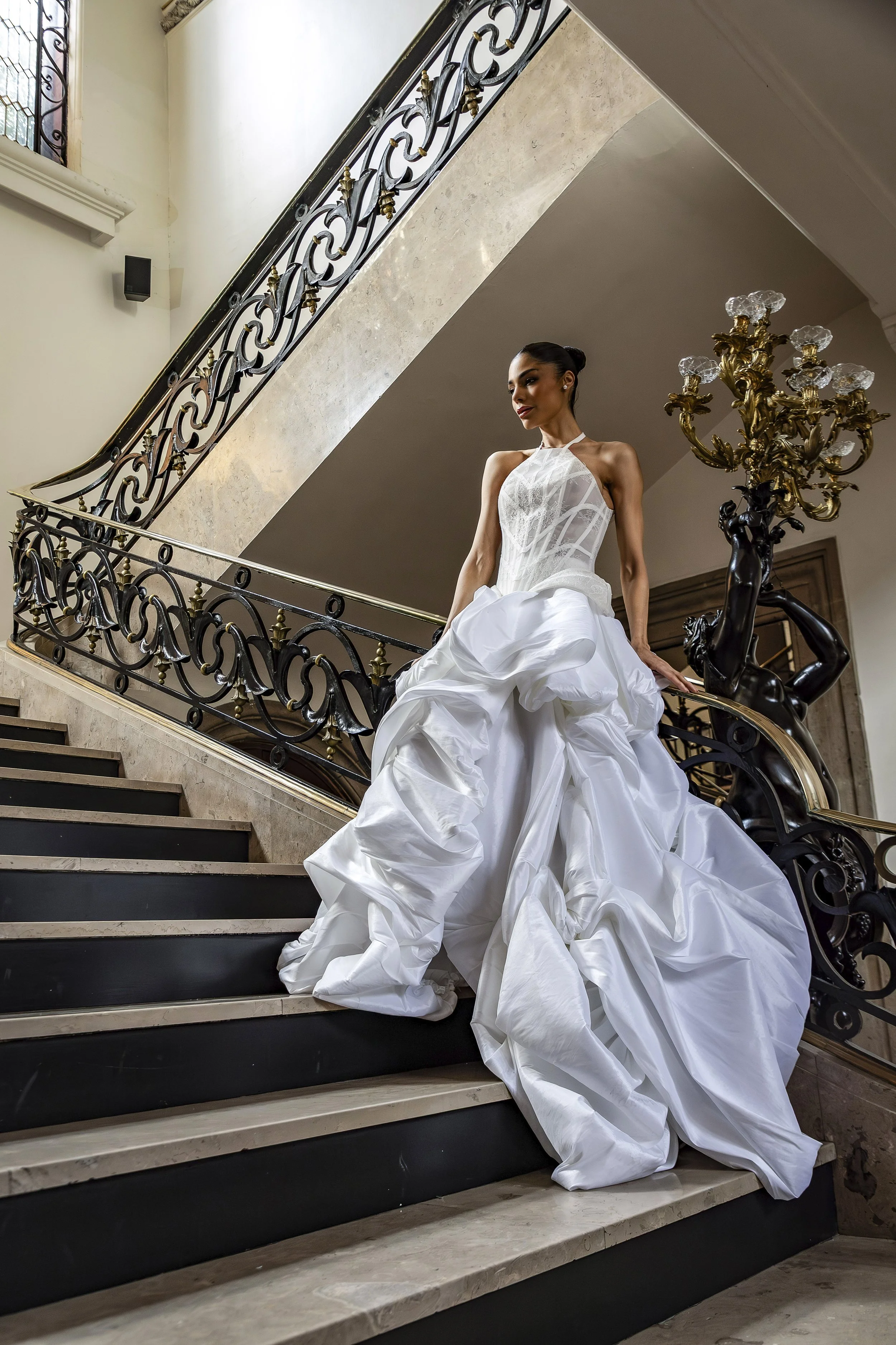 A woman in a white wedding gown sitting on a staircase with ornate black and gold railing, a chandelier, and large windows in the background.
