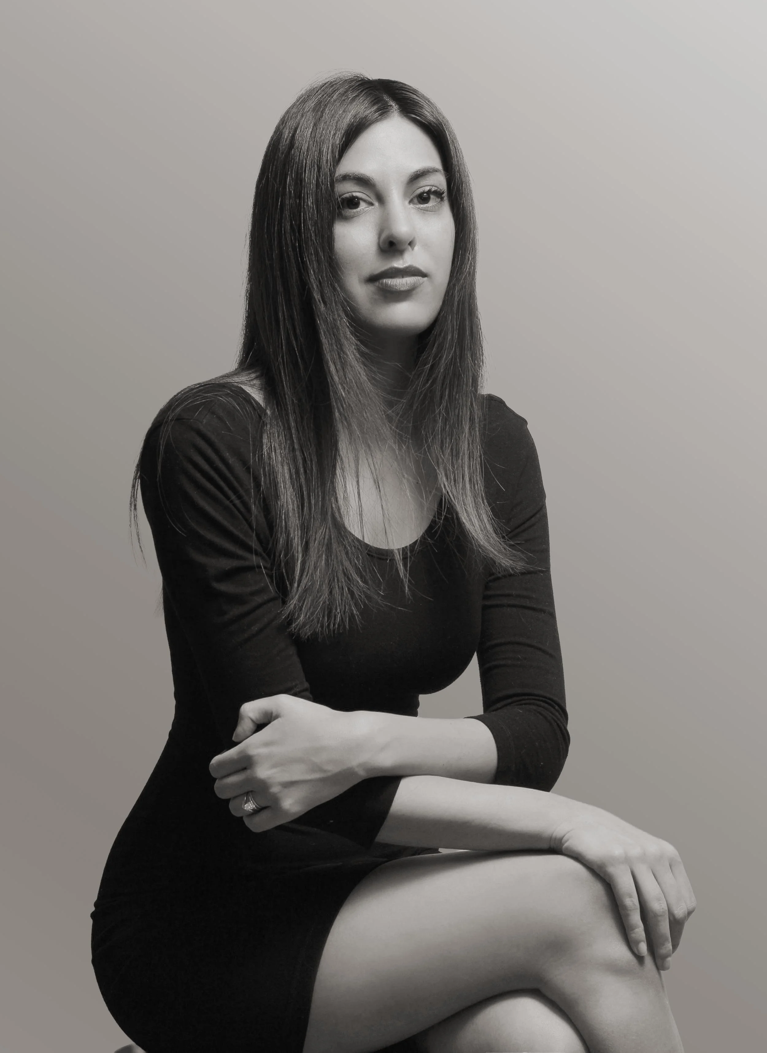 A black and white photo of a woman with long, straight hair, wearing a form-fitting black dress, seated with her arms crossed, looking at the camera with a neutral expression, against a plain light background.