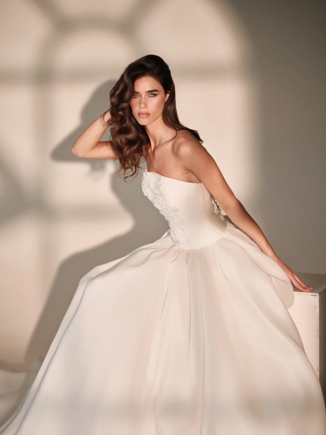 Maria Pia at New York Bridal Market.  A woman with long, wavy brown hair in a strapless white dress sitting on a white block with shadows cast on a neutral background.