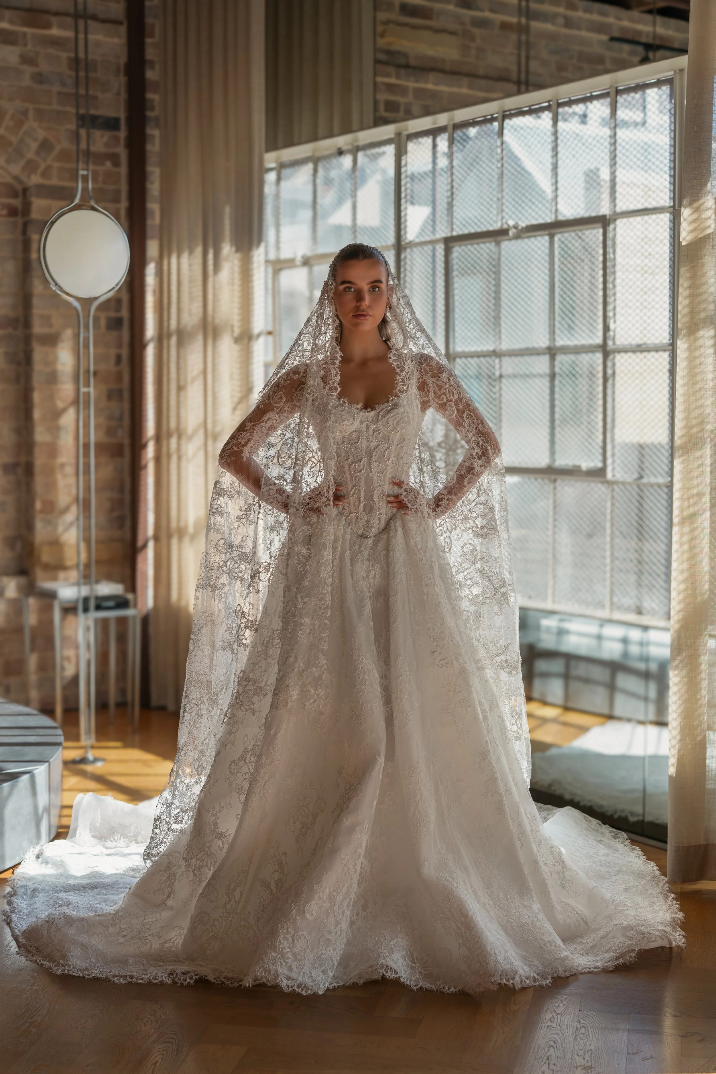 Blanche Bridal at New York Bridal Market  A woman wearing a wedding dress with lace details, standing in a room with large windows, brick walls, and curtain panels, illuminated by natural light.