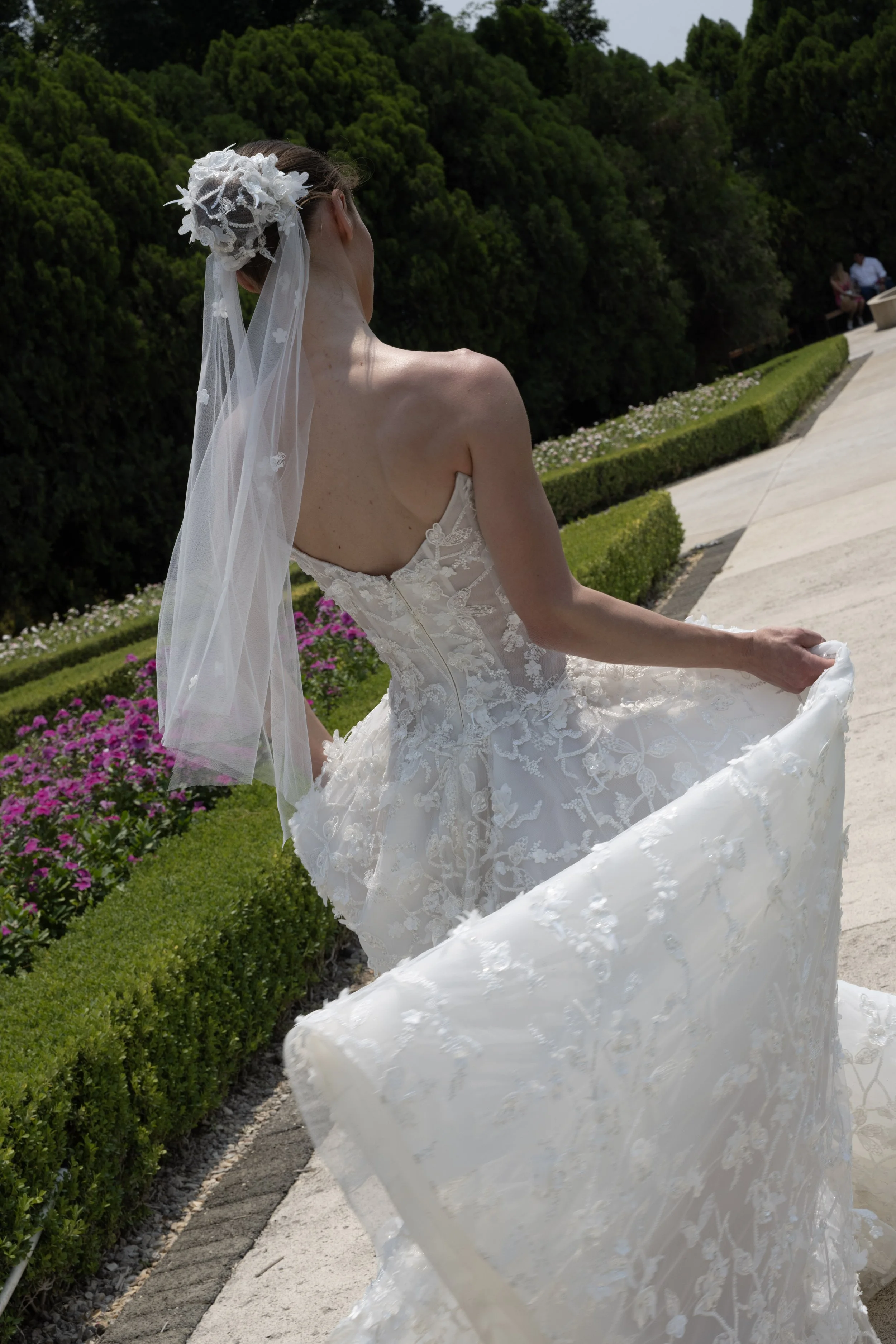Guardiola  at New York Bridal Market.  A bride holding up her wedding dress outdoors with greenery and flowers in the background.