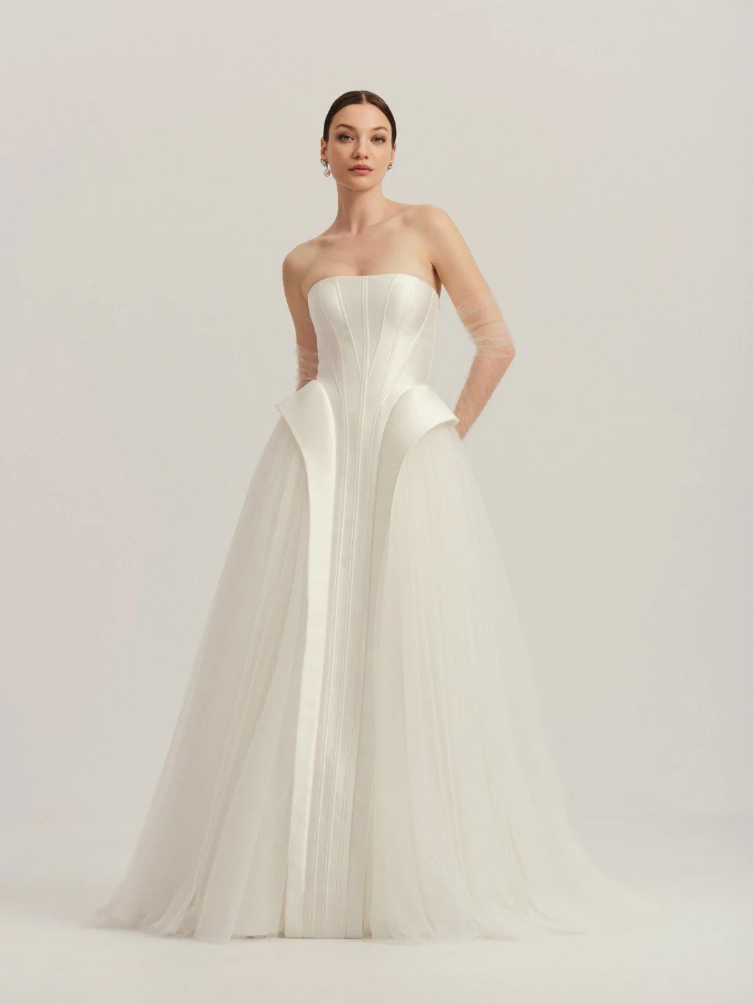 Divine Atelier at New York Bridal Maket.  Woman in a strapless white wedding gown with sheer long sleeves, standing against a plain neutral background.