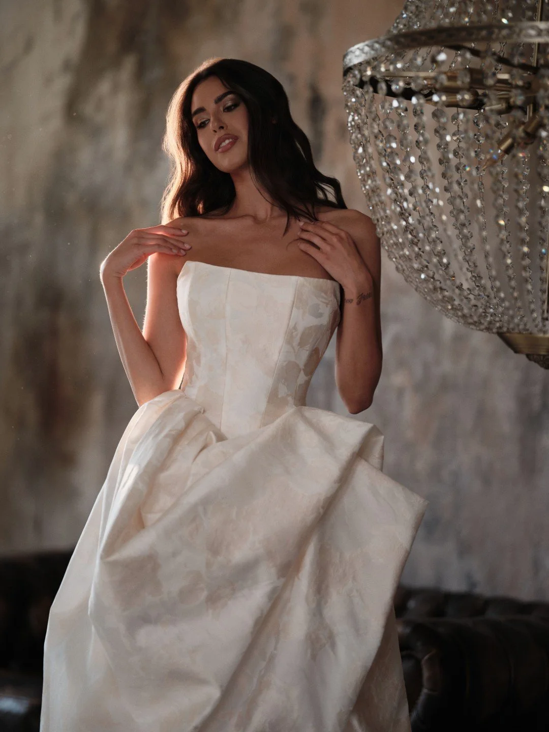 Maria Pia at New York Bridal Market A woman with dark hair wearing a strapless white gown with a textured pattern, standing near a large, ornate crystal chandelier, in front of a rustic wall.