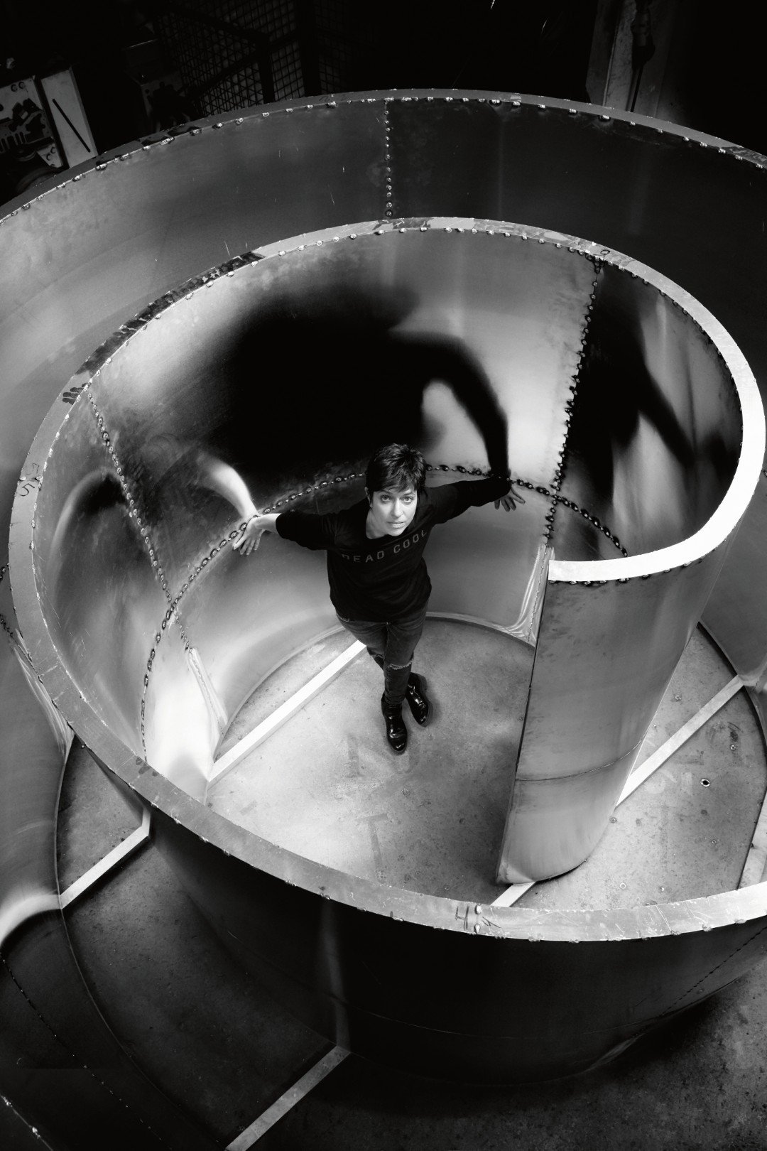 A person standing inside a large metal spiral staircase, looking up, with shadows cast on the inner wall of the staircase.