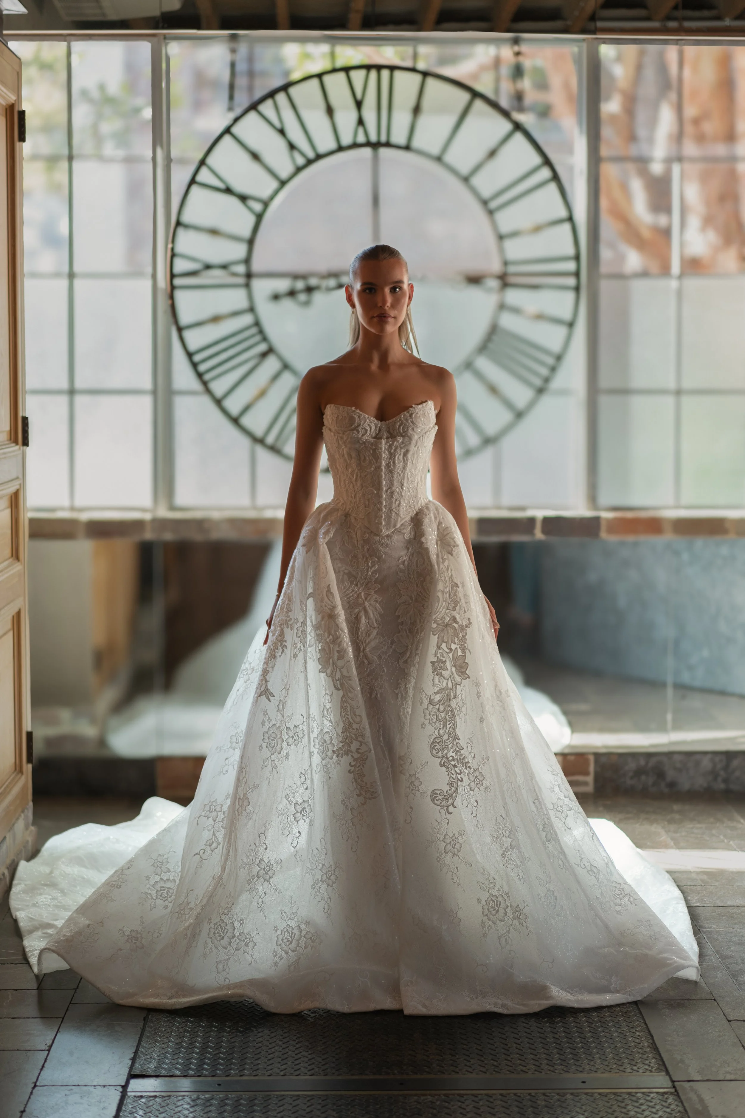 Blanche Bridal at New York Bridal Market  A woman in a wedding dress standing in front of a large clock inside a building with glass windows.