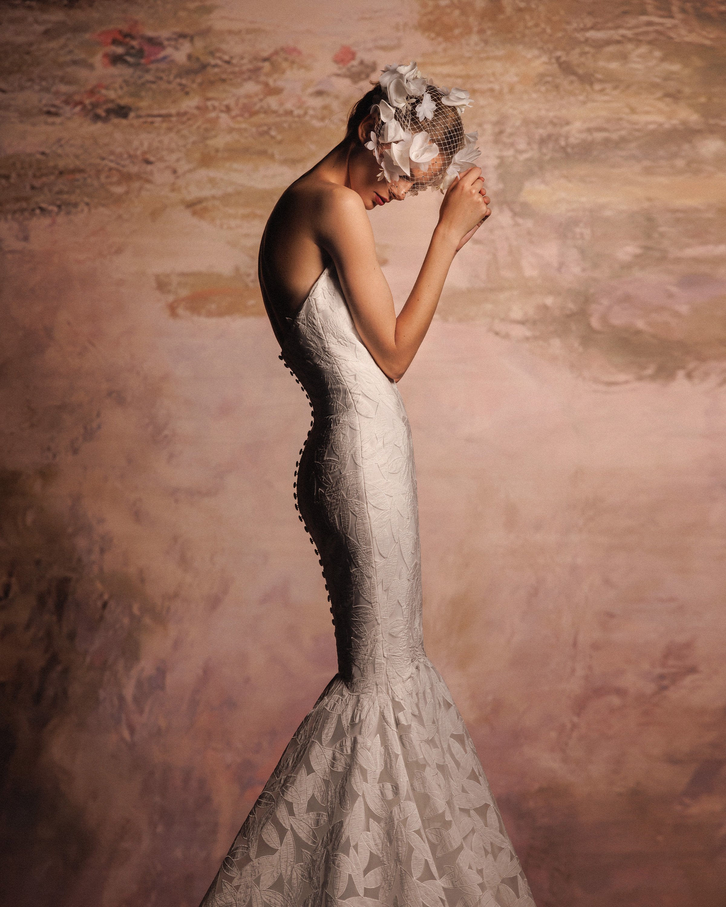 Gemy Maalouf at New York Bridal Makrket A woman wearing a strapless white dress with lace detail, elegant makeup, and a floral headpiece with netting, standing against a painted backdrop.