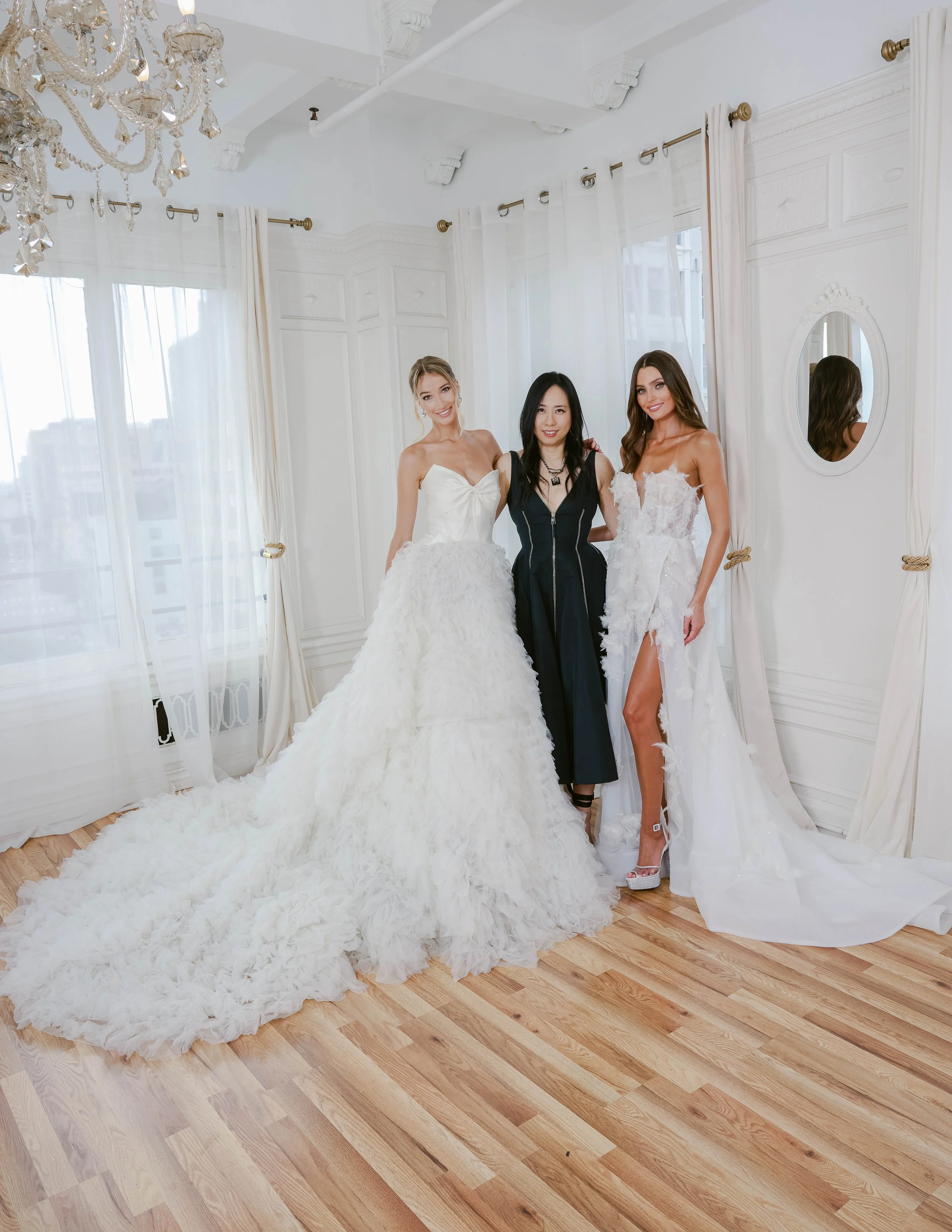 Three women standing in a bright, elegant room with white walls, curtains, and a chandelier; two women are dressed in bridal gowns, and one in black attire.