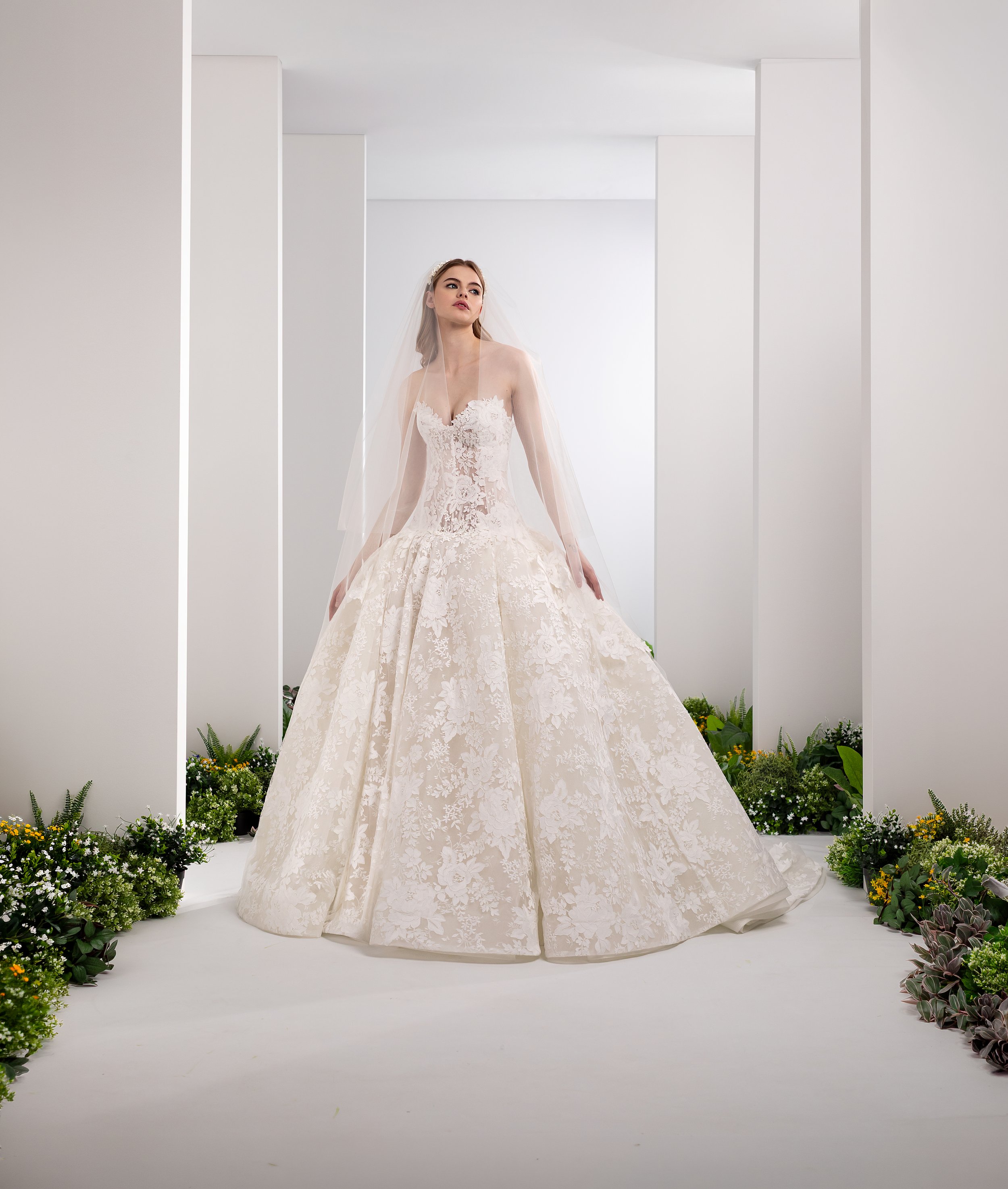 Valentini Spose at New York Bridal Market.   A bride wearing a white lace wedding gown with floral embroidery, standing in a minimalist space with white walls and surrounded by plants and flowers.