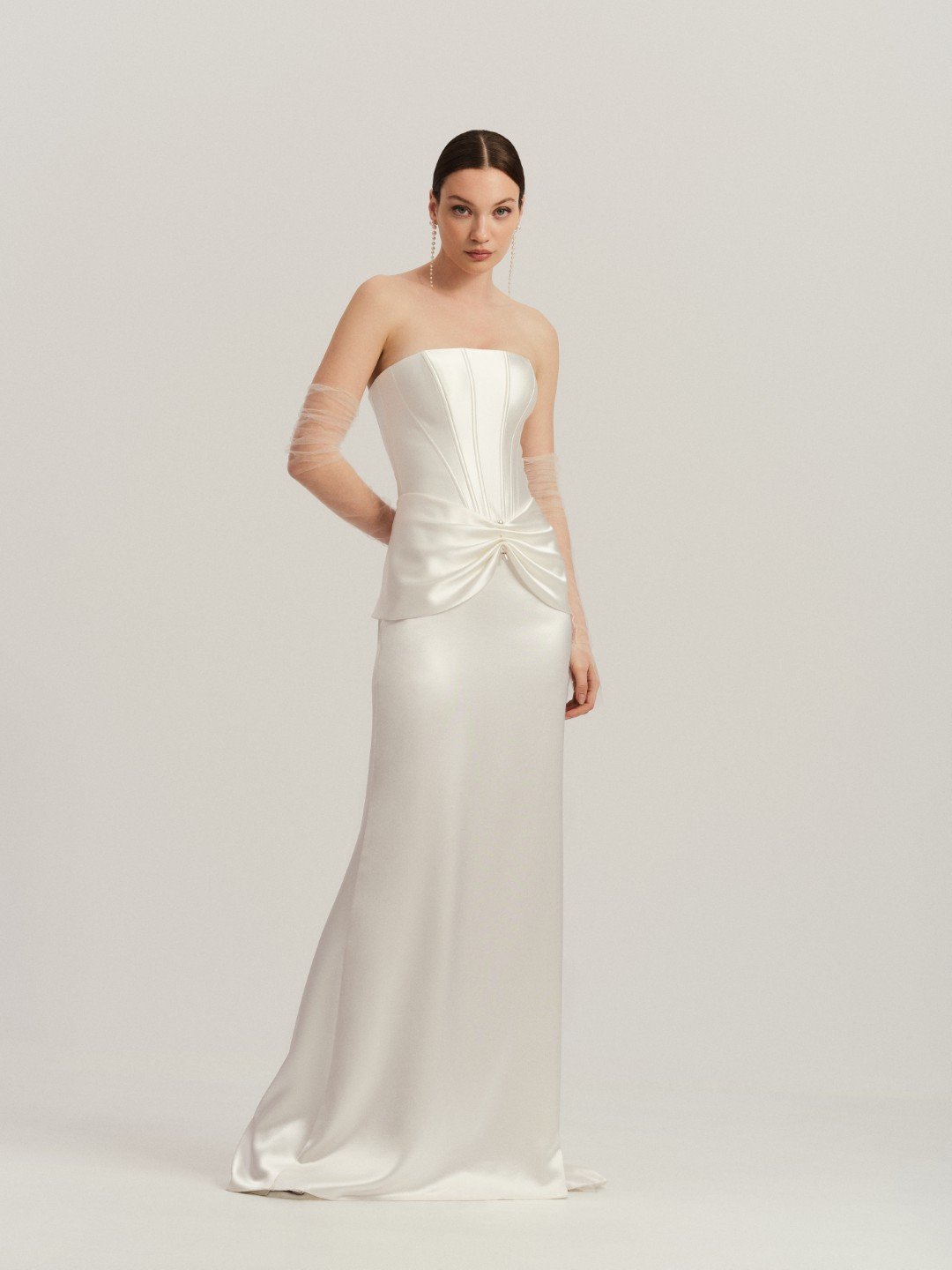 Divine Atelier at New York Bridal Maket.  Woman in a strapless white satin wedding gown with long sheer sleeves, standing against a plain light background.