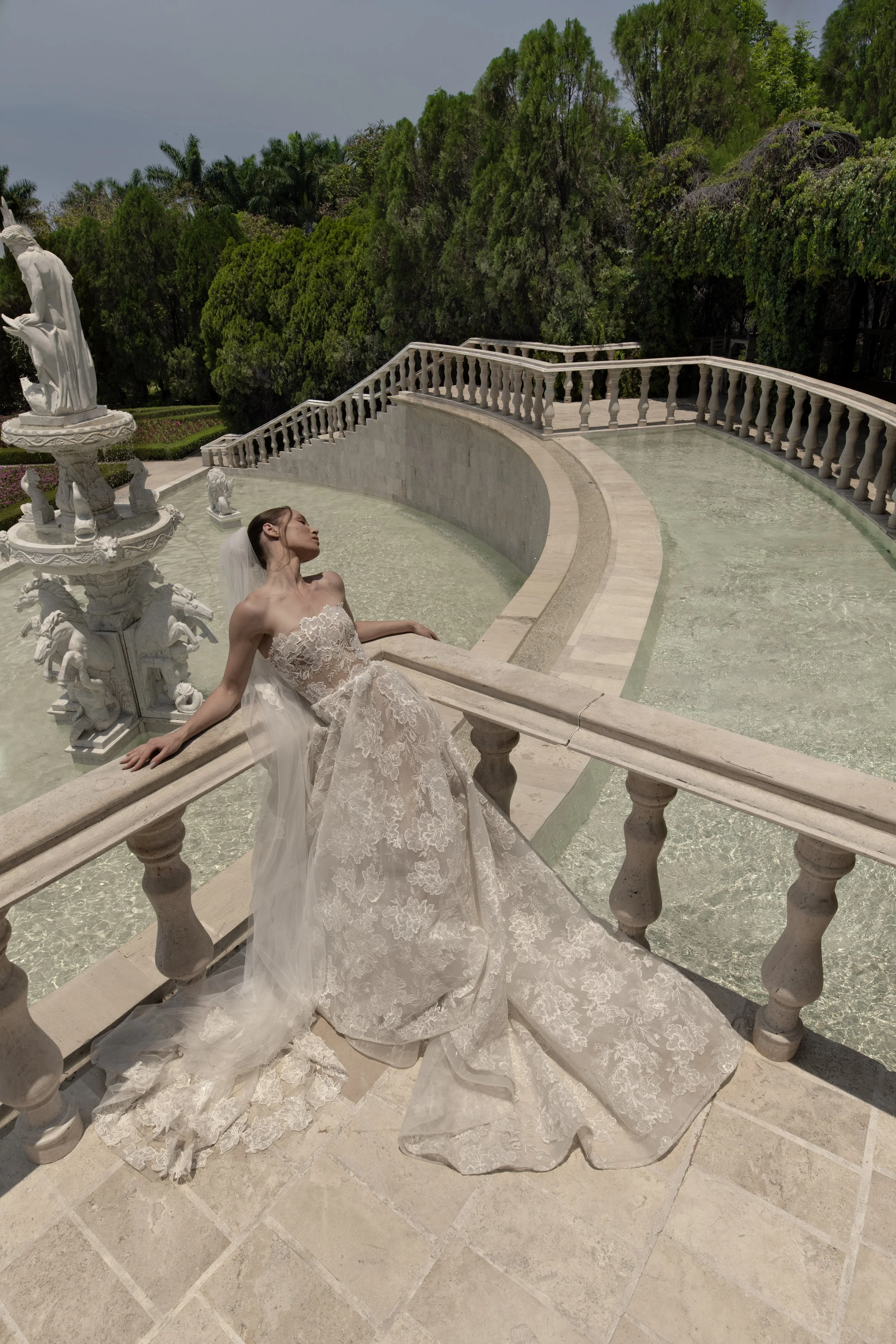 A woman in a white lace wedding dress with a long train and veil is reclining on a stone balustrade next to a water fountain with a statue, in a garden setting with lush green trees in the background.