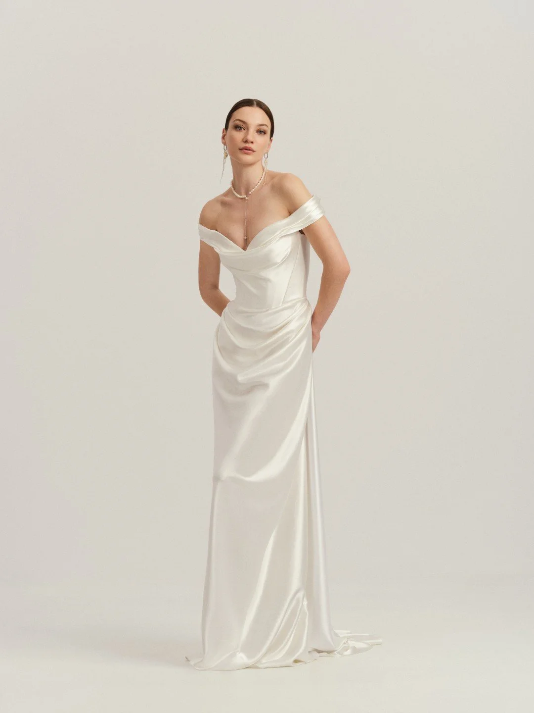 Divine Atelier at New York Bridal Maket. Woman in a white satin off-shoulder dress with pearl jewelry posing against a plain light background.