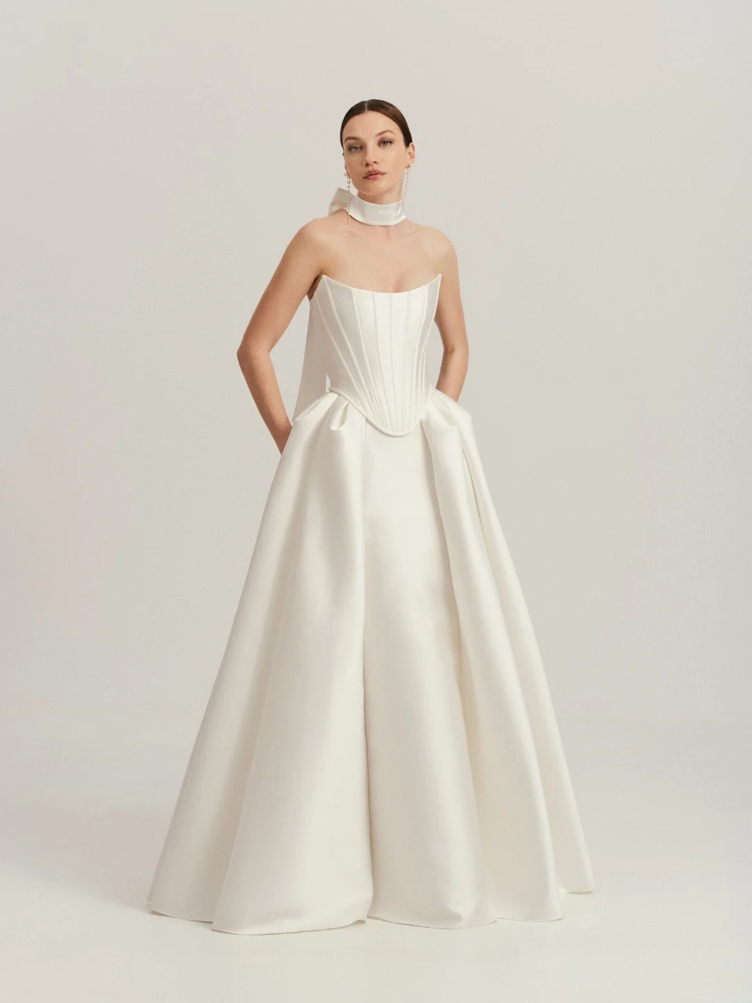 Divine Atelier at New York Bridal Maket.  Woman in a strapless, white, floor-length gown with a fitted bodice and full skirt, standing against a plain background.