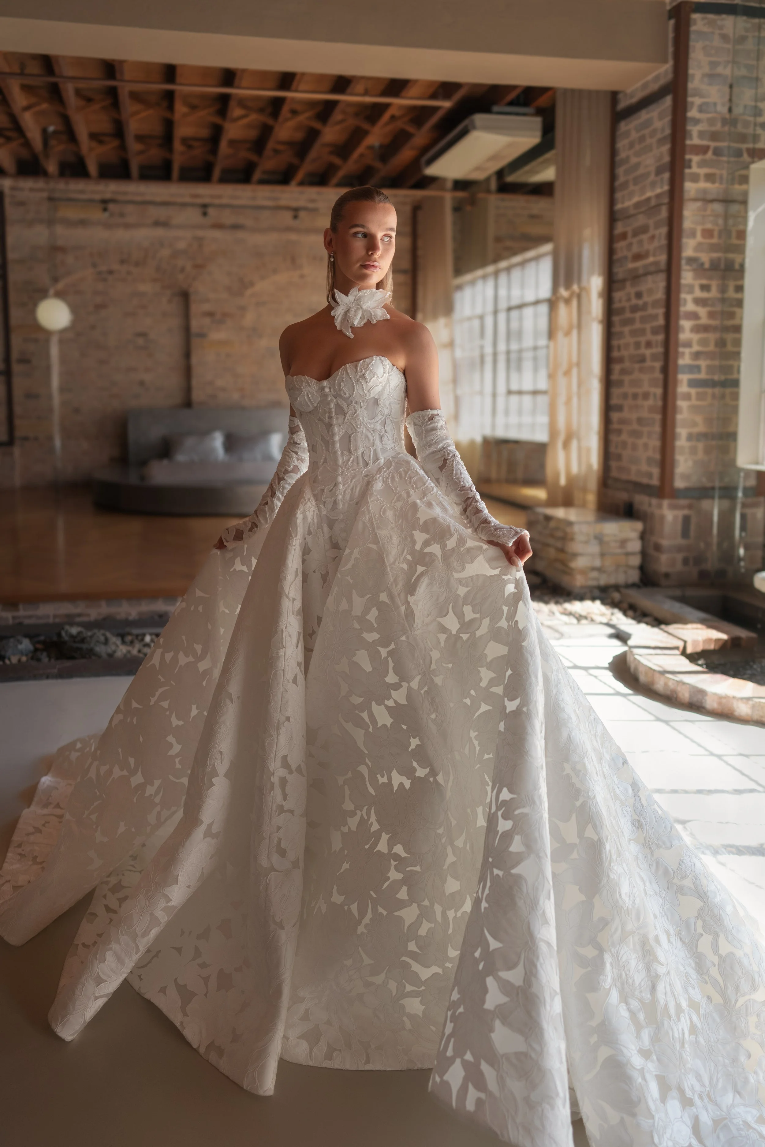 Blanche Bridal at New York Bridal Market  A woman in a white, strapless wedding gown with lace overlay and floral patterns, standing in a bright, brick-walled room with large windows and curtains.
