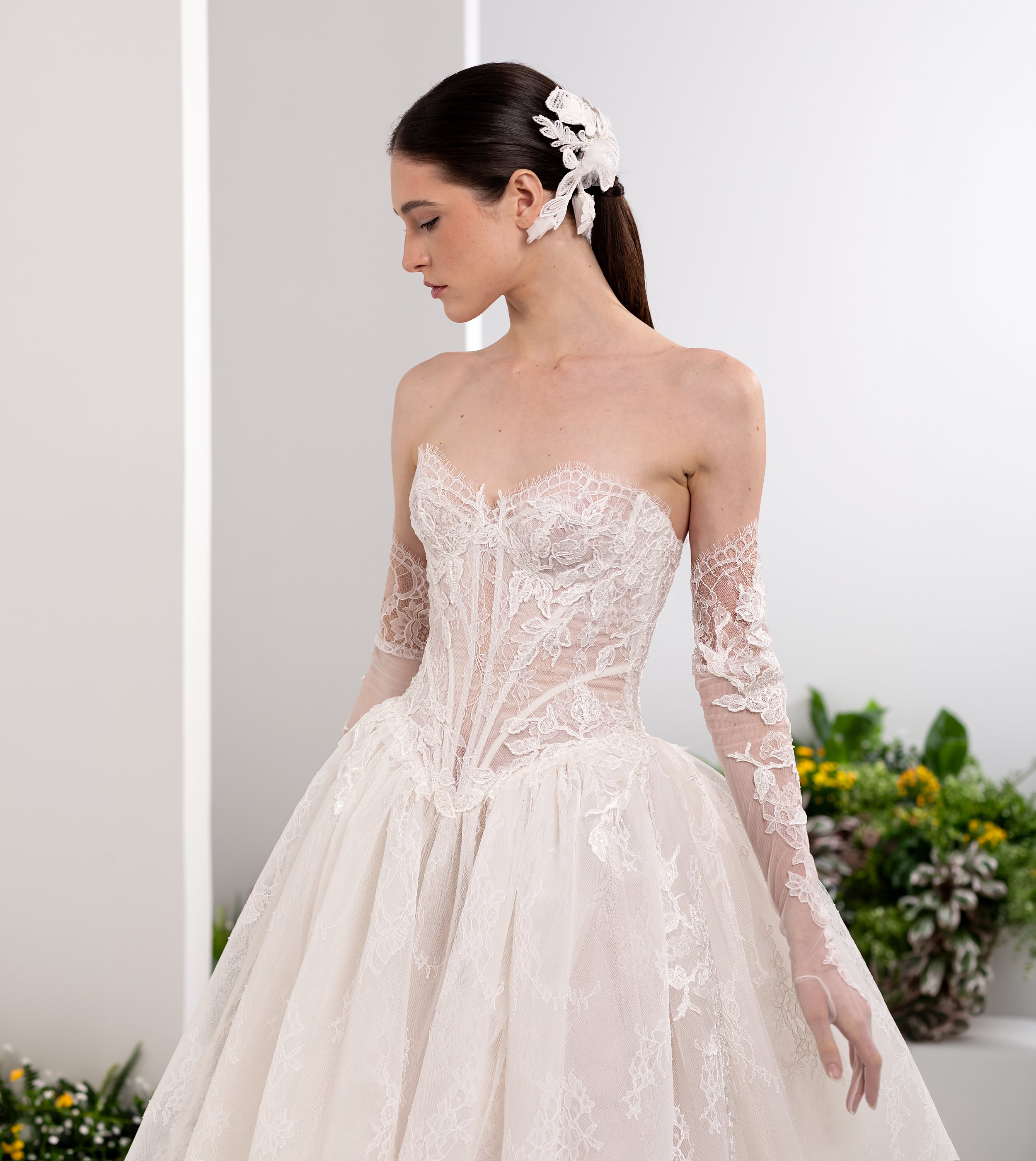 Valentini Spose at New York Bridal Market.  A woman with dark hair styled in a ponytail, adorned with a lace hair accessory, wearing a delicate lace wedding dress with long lace sleeves, standing outdoors with flowers in the background.