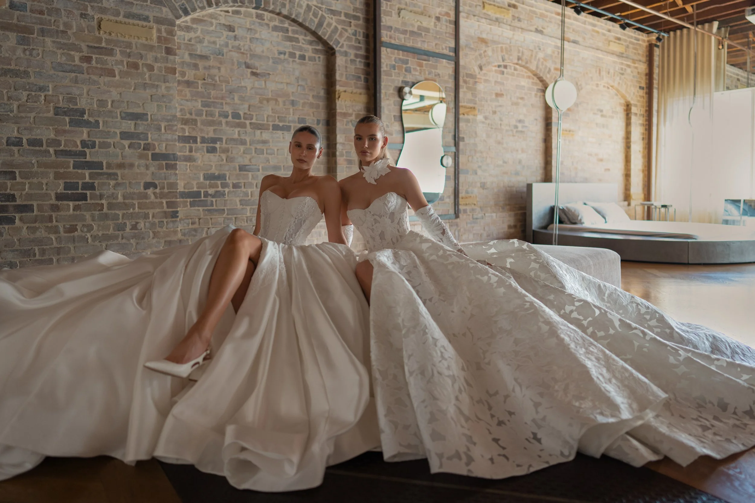 Blanche Bridal at New York Bridal Market Two women in wedding dresses sit on a bed in a modern, industrial-style room with exposed brick walls and large windows. They are facing the camera with neutral expressions.