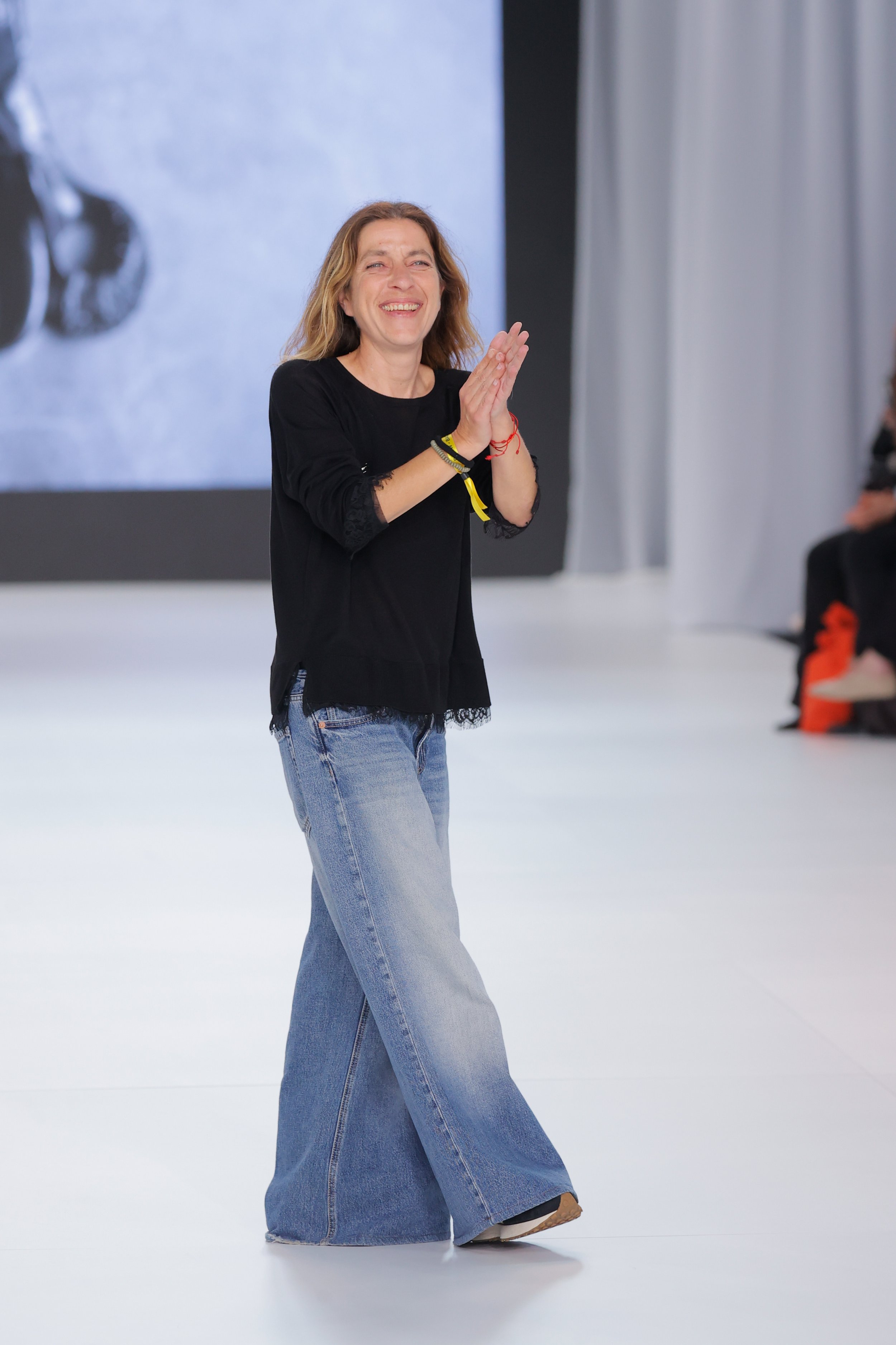 Yolancris at New York Bridal Market A woman in casual clothing, smiling and clapping, in a studio with a large abstract background.