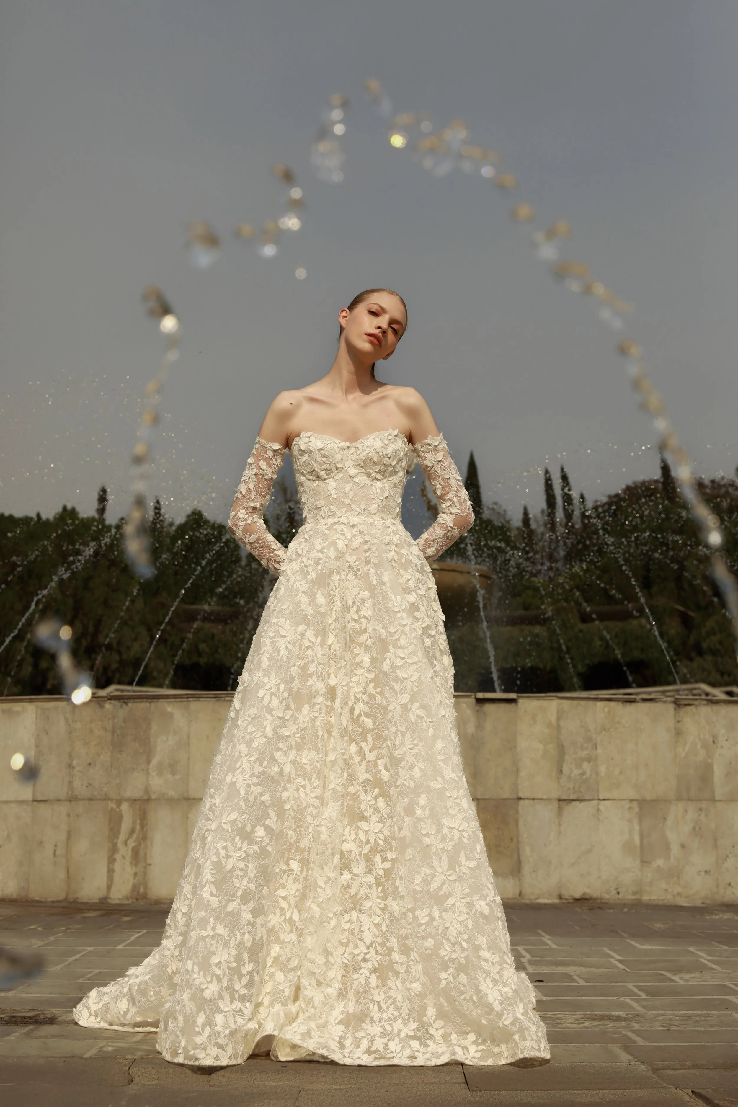 	
Guardiola at New York Bridal Market A woman in a white lace wedding dress stands in front of a fountain with water arcing from it, with trees and a cloudy sky in the background.