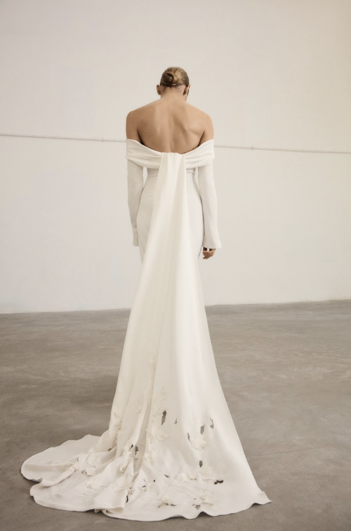 Marmar Halim at New York Bridal Market A person wearing an elegant, off-the-shoulder white gown with a long train stained and damaged at the bottom, standing with their back facing the camera in a plain, empty room.