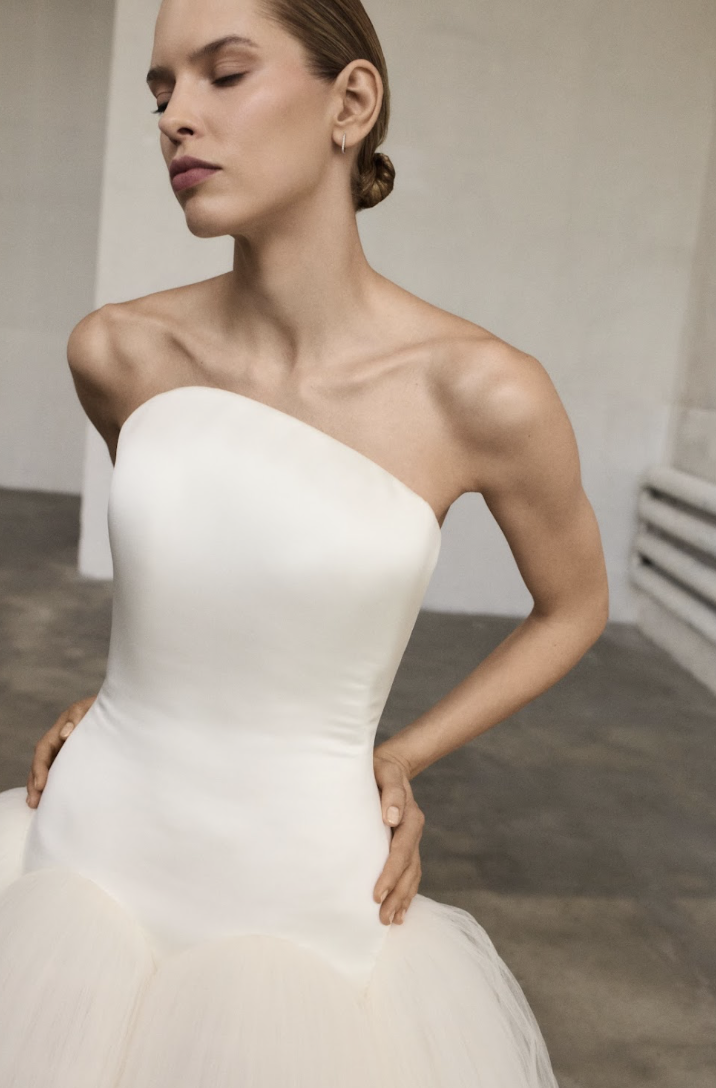 	
Marmar Halim at New York Bridal Market A woman in a strapless white wedding dress with a fitted bodice and full skirt stands with her hands on her hips, with her eyes closed and head slightly turned to the side, in an indoor setting.