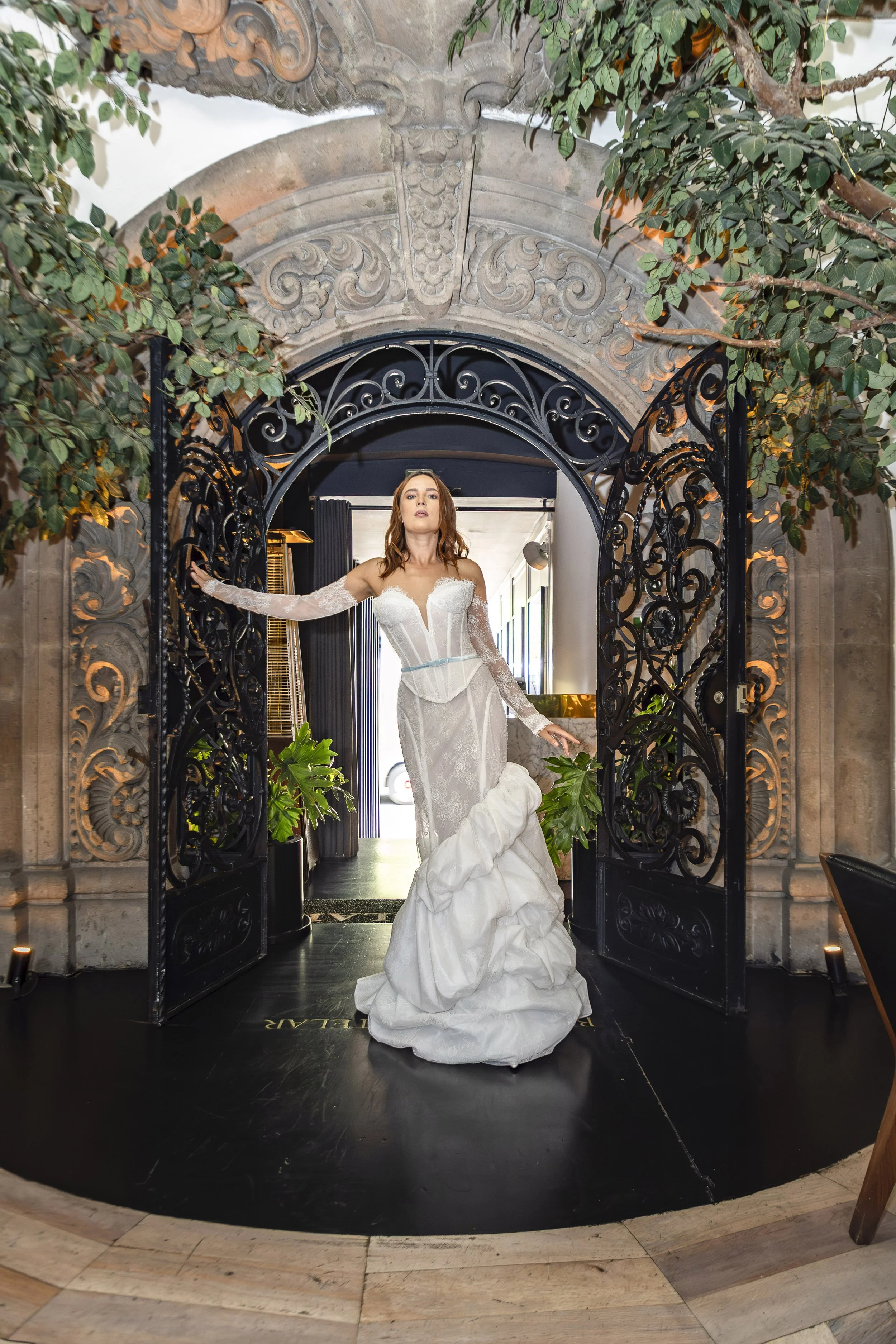A woman in an elegant white wedding gown standing in an ornate doorway with black wrought iron gates, surrounded by lush greenery and decorative stone arch and carvings.