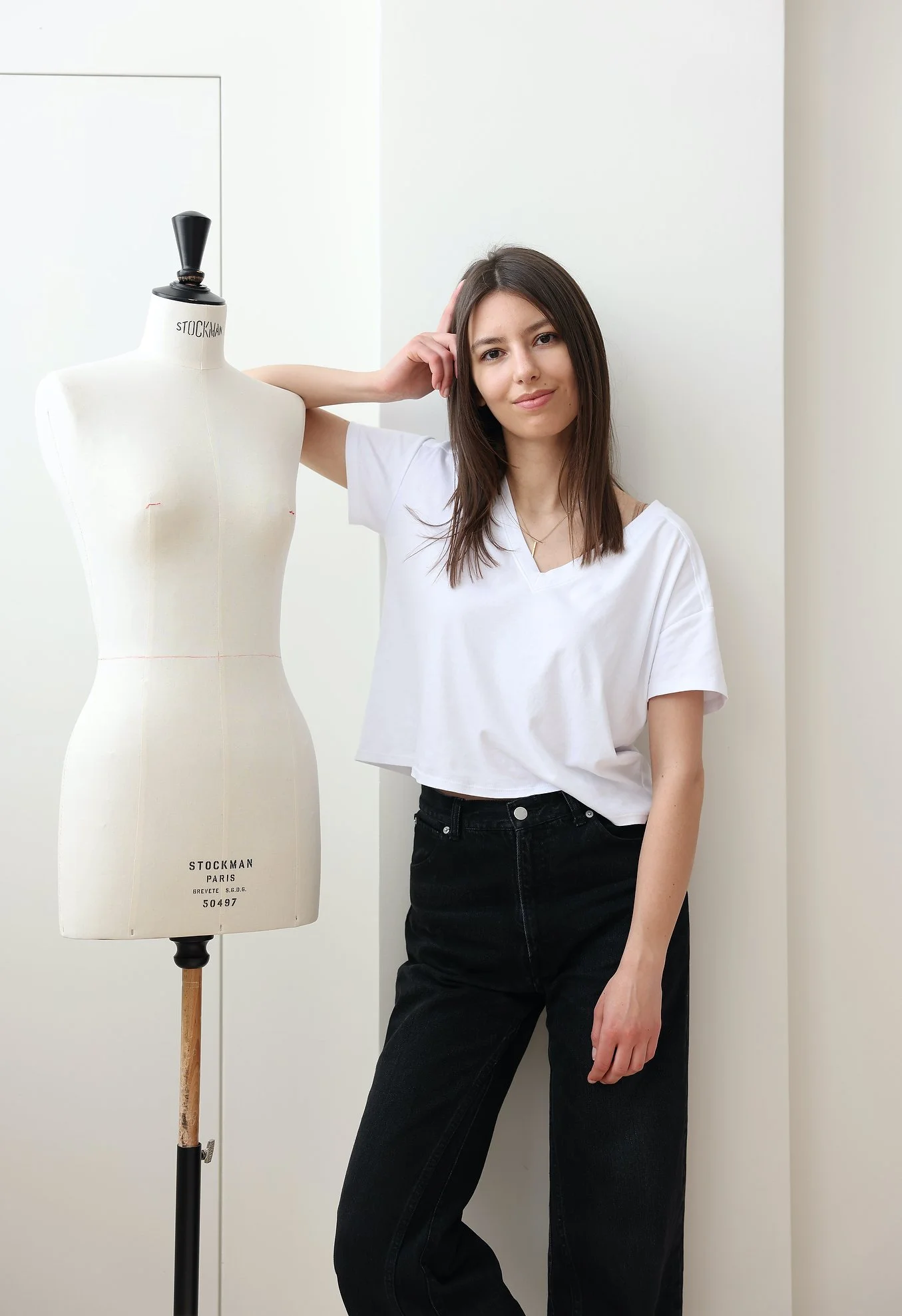 Divine Atelier at New York Bridal Market. Young woman standing next to a dress form mannequin, posing casually with her hand on her head in a bright, plain room.