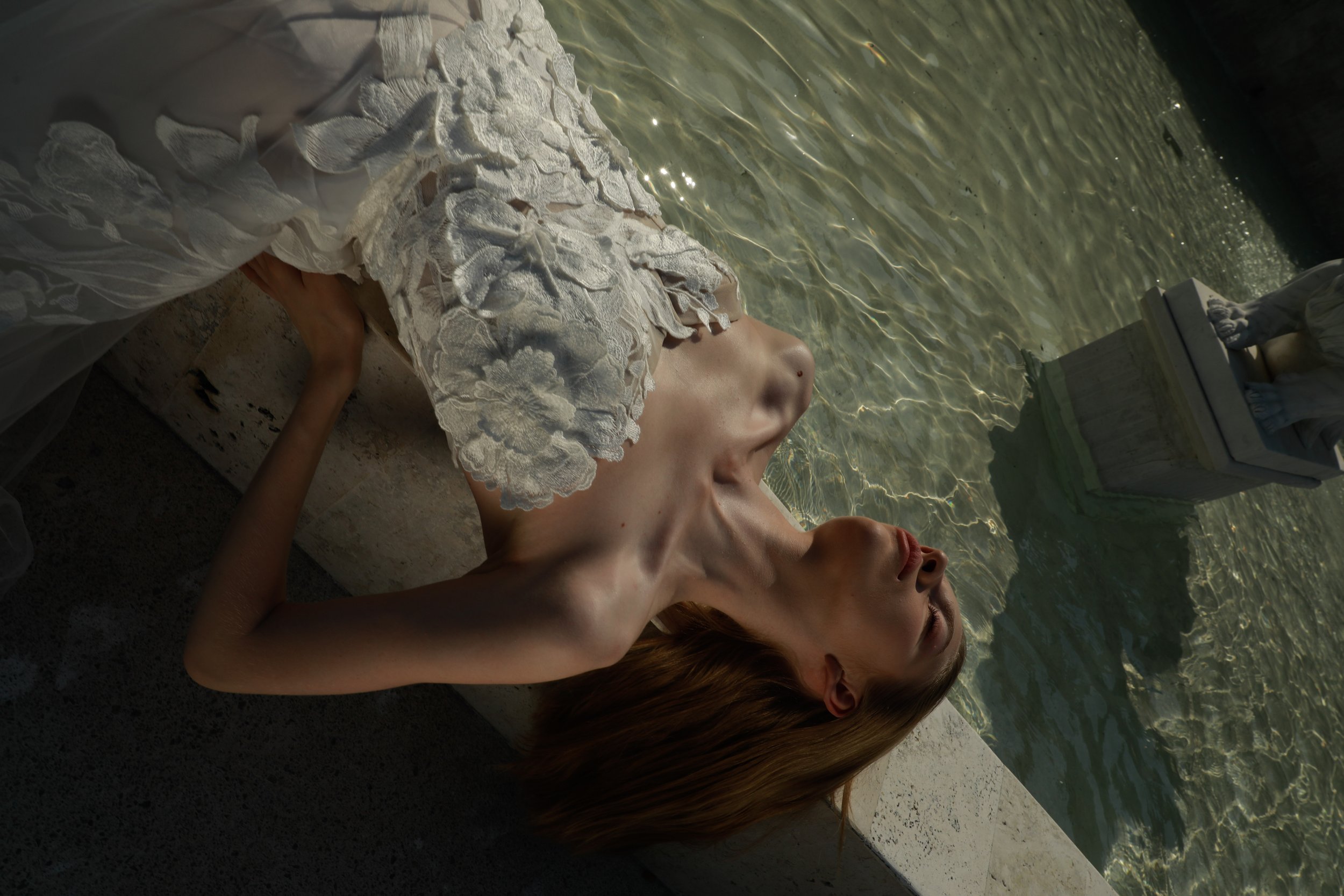 	
Guardiola at New York Bridal Market A person with red hair and light skin lying in water, partially covered by a white lace dress, with their head resting on the edge of a pool or body of water. The water has sunlight reflections.