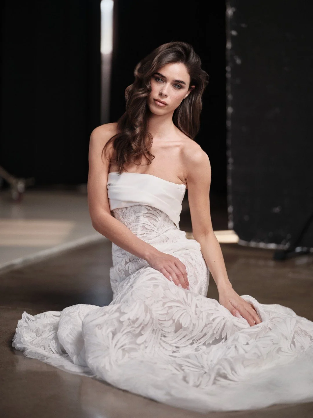 Maria Pia at New York Bridal Market.  A woman with long, wavy brown hair and blue eyes is sitting on the floor, wearing a strapless white dress with lace detailing, in a studio setting with black and white backgrounds.