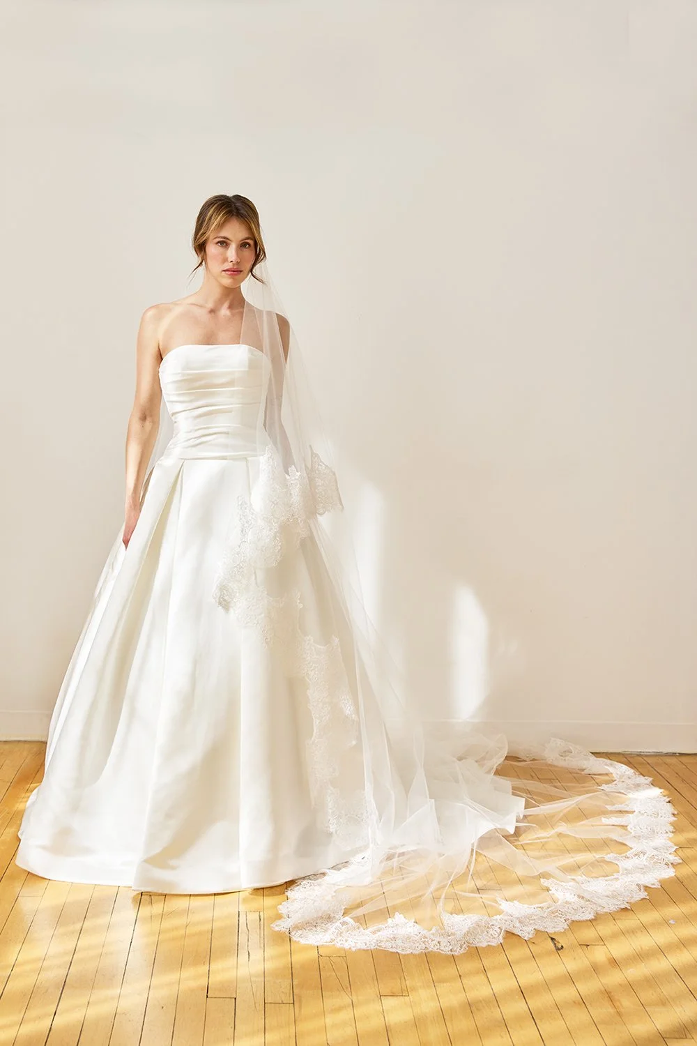 A woman wearing a white wedding dress with a strapless bodice and lace shoulder detail, standing on a hardwood floor against a plain beige wall. She has a veil and the dress has a long train with lace trim.