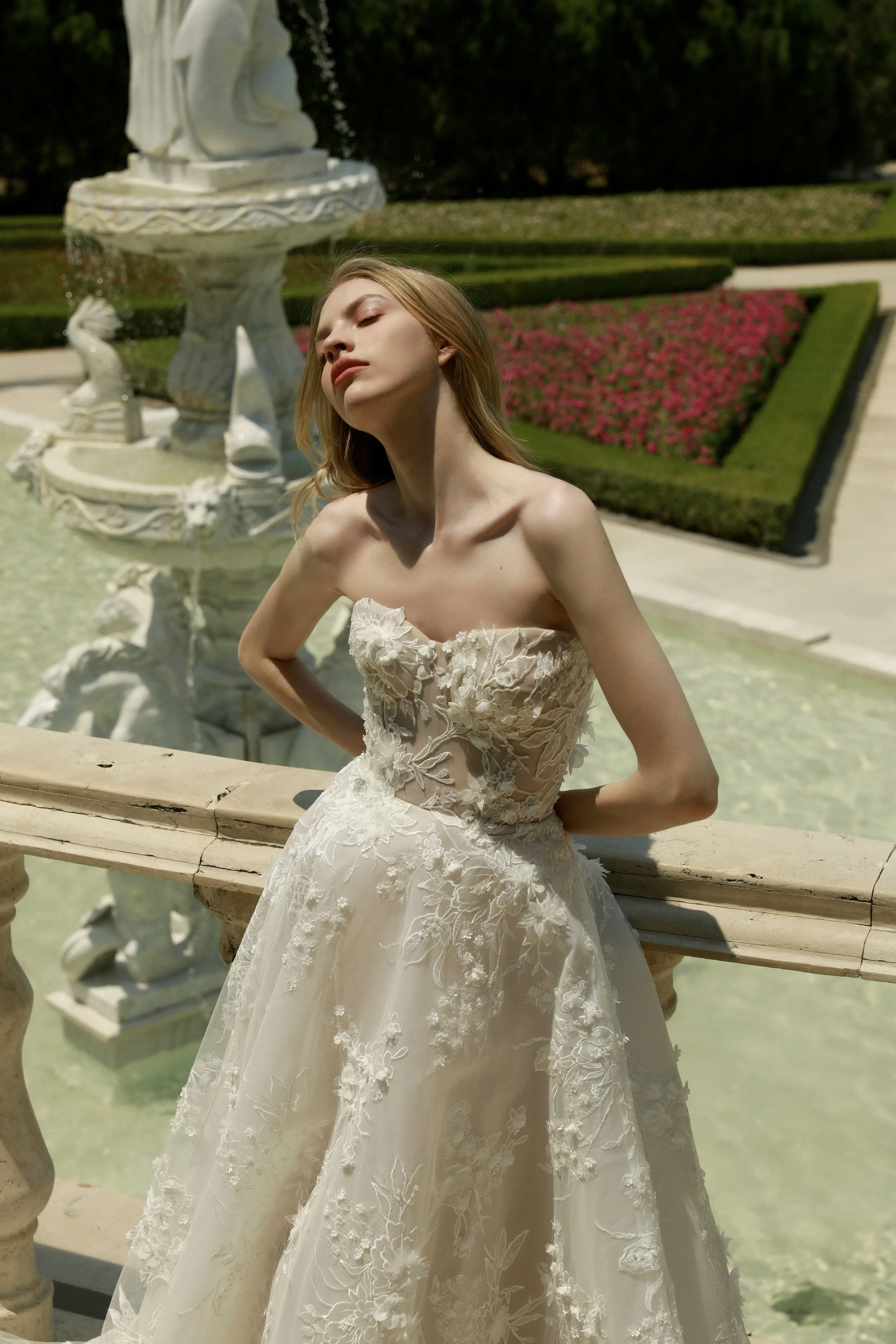 A woman in a wedding dress standing outdoors near a fountain with a garden and pink flowers in the background.
