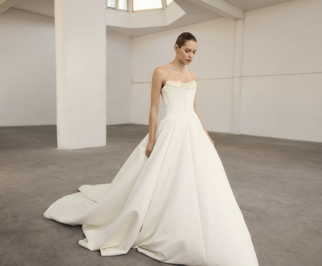 A woman in a white wedding gown standing in a minimalist, industrial space with gray concrete floors and white walls.