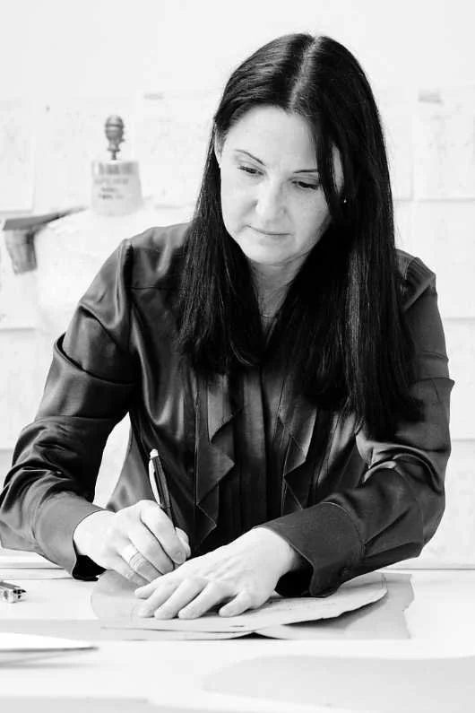 Black and white photo of a woman with dark hair working on a craft project at a table, surrounded by papers and tools, in an indoor setting with papers on the wall behind her.