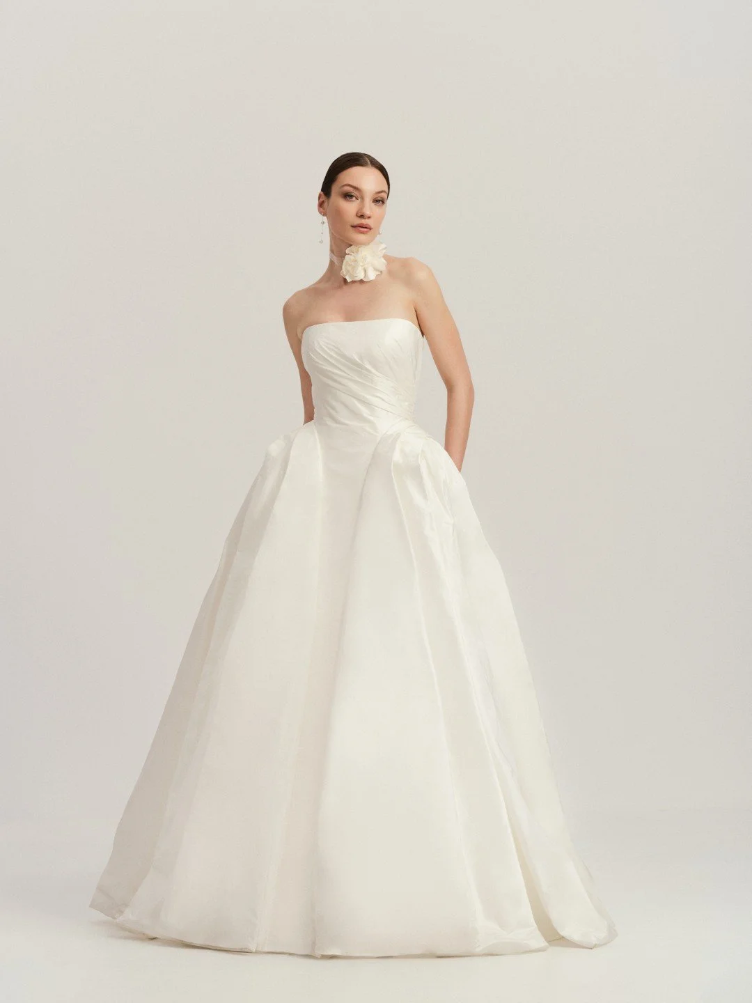 Divine Atelier at New York Bridal Market A woman in a strapless white wedding gown with a full skirt, standing against a plain light background, wearing a large white flower choker and earrings.