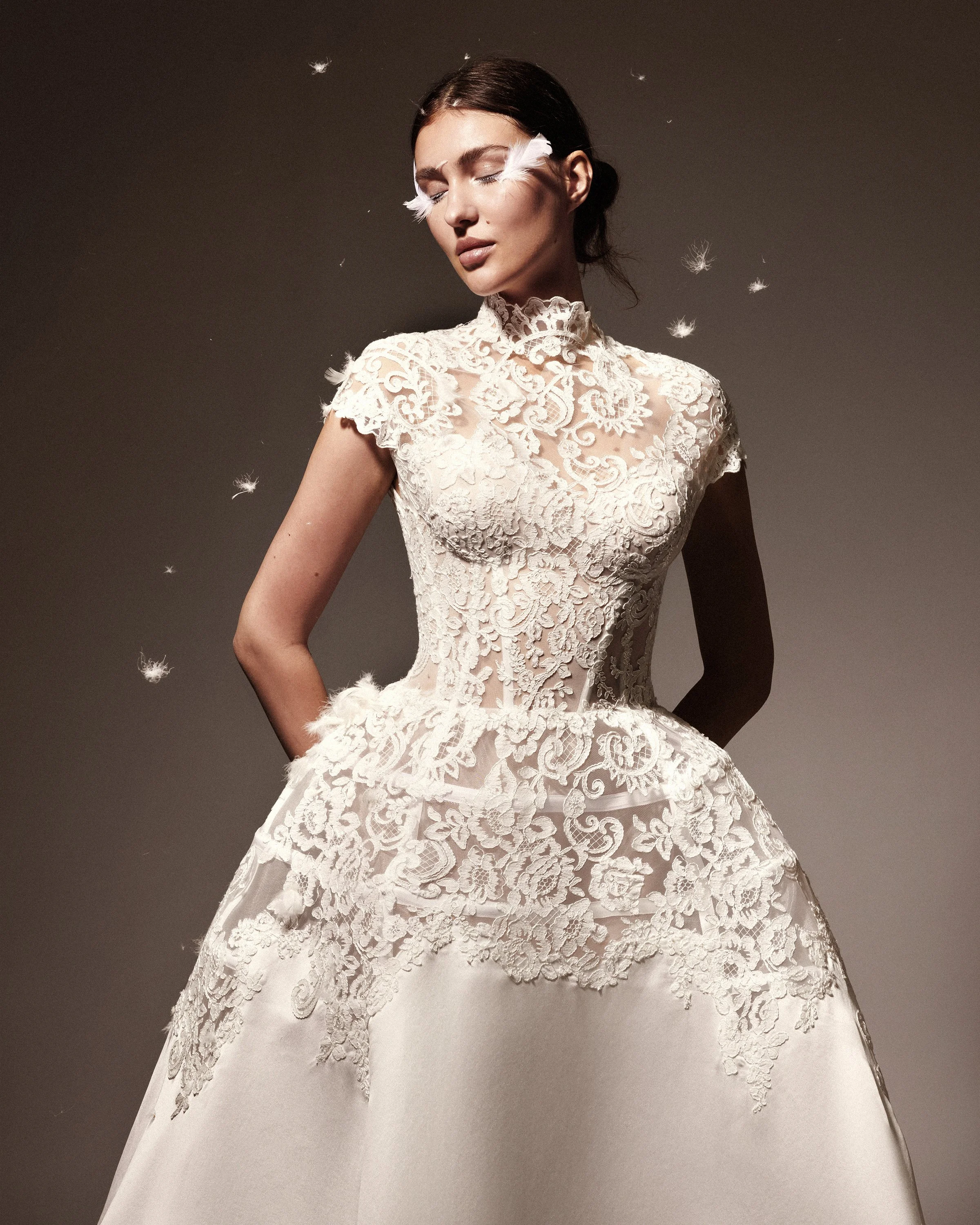 Gemy Maalouf at New York Bridal Makrket A woman in a white, lace wedding dress with a high neckline, closed eyes, and dark hair, stands against a plain dark background with floating white feathers.