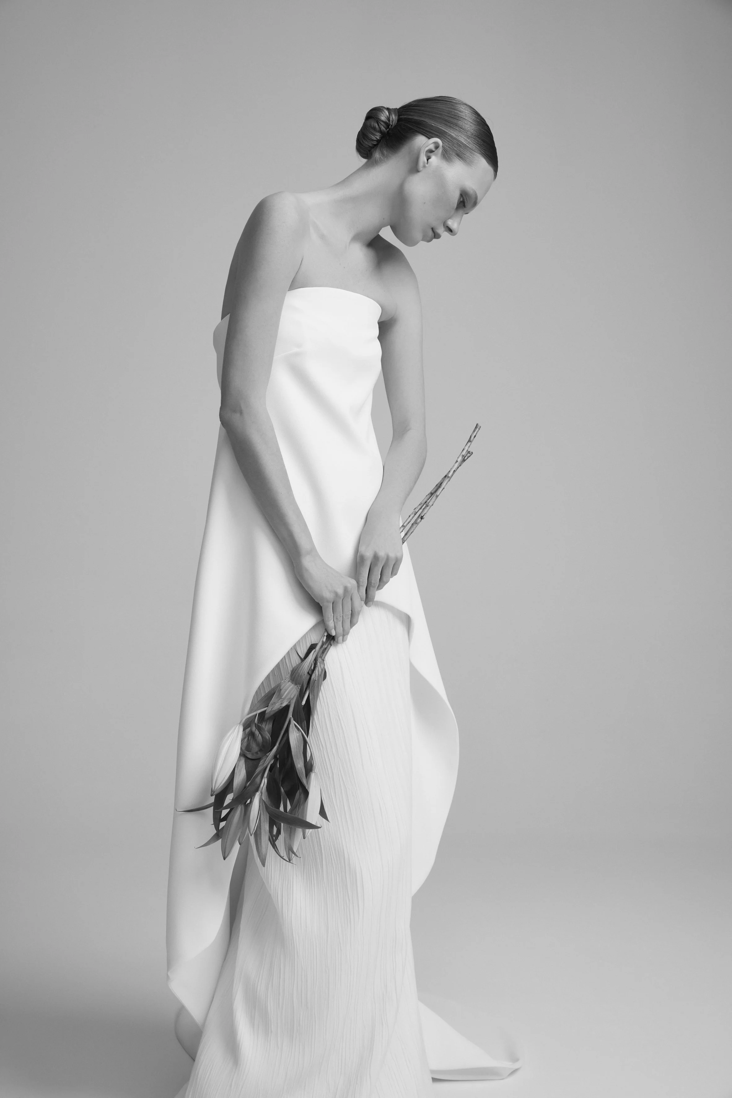 Sophie et Voila at New York Bridal Market. A black-and-white photo of a woman in an elegant, strapless gown holding a bunch of leaves and what appears to be a wooden stick, with her head bowed and hair styled in an updo.