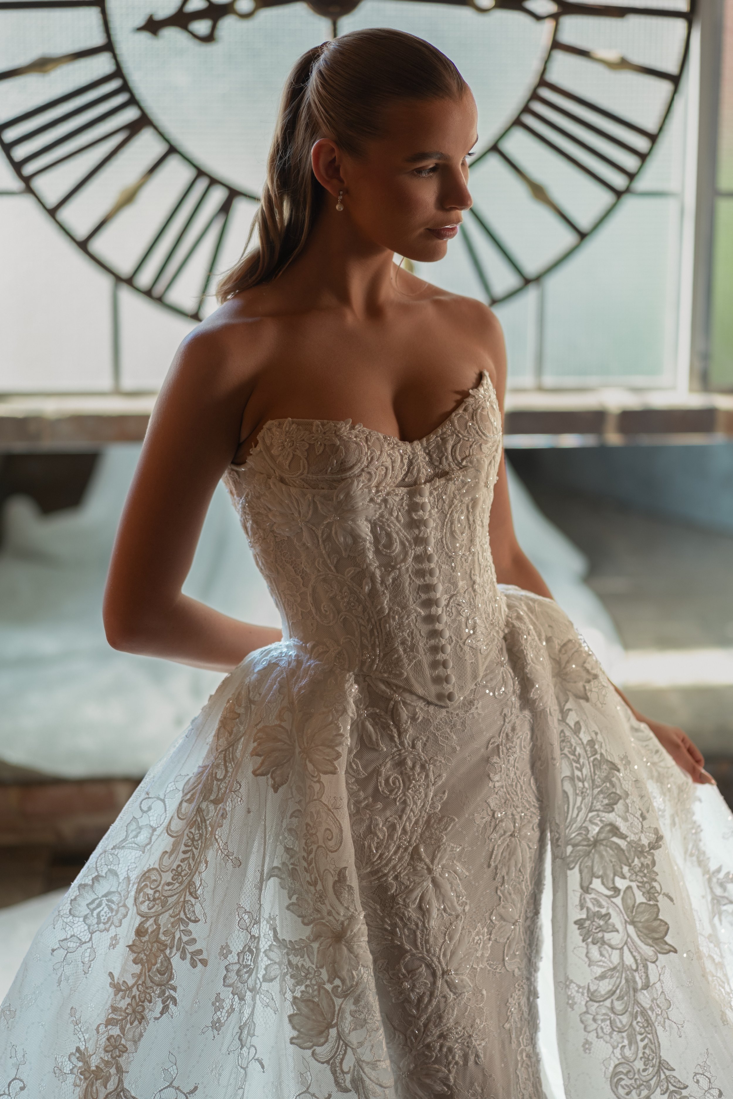 Blanche Bridal at New York Bridal Market  A woman in an elegant off-shoulder wedding dress with intricate lace floral embroidery, standing indoors near a large clock window, with her hair styled long and straight, and wearing pearl earrings.