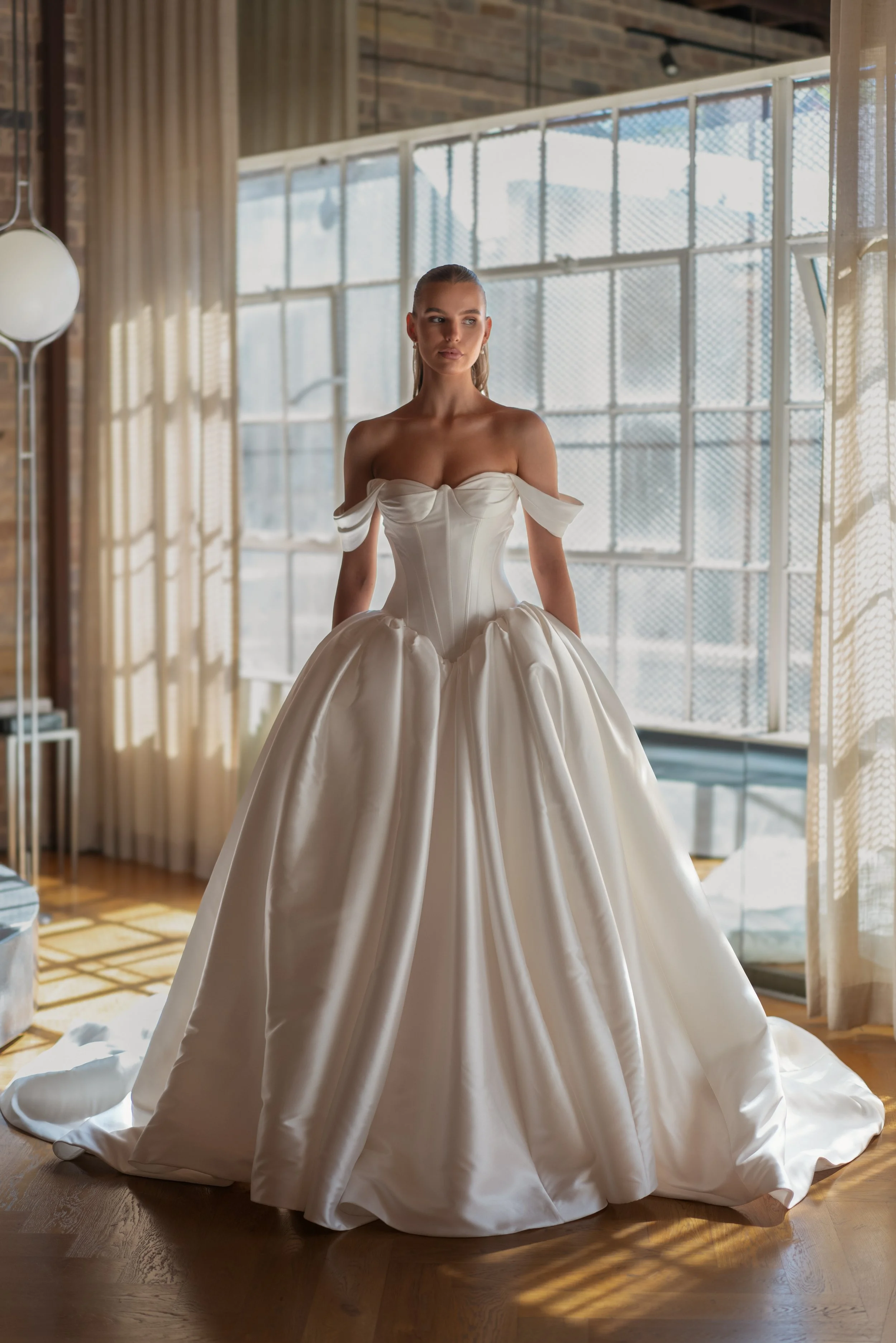 Blanche Bridal at New York Bridal Market  A woman in a strapless white wedding gown with off-the-shoulder sleeves, standing indoors in front of large windows with curtains and natural light.