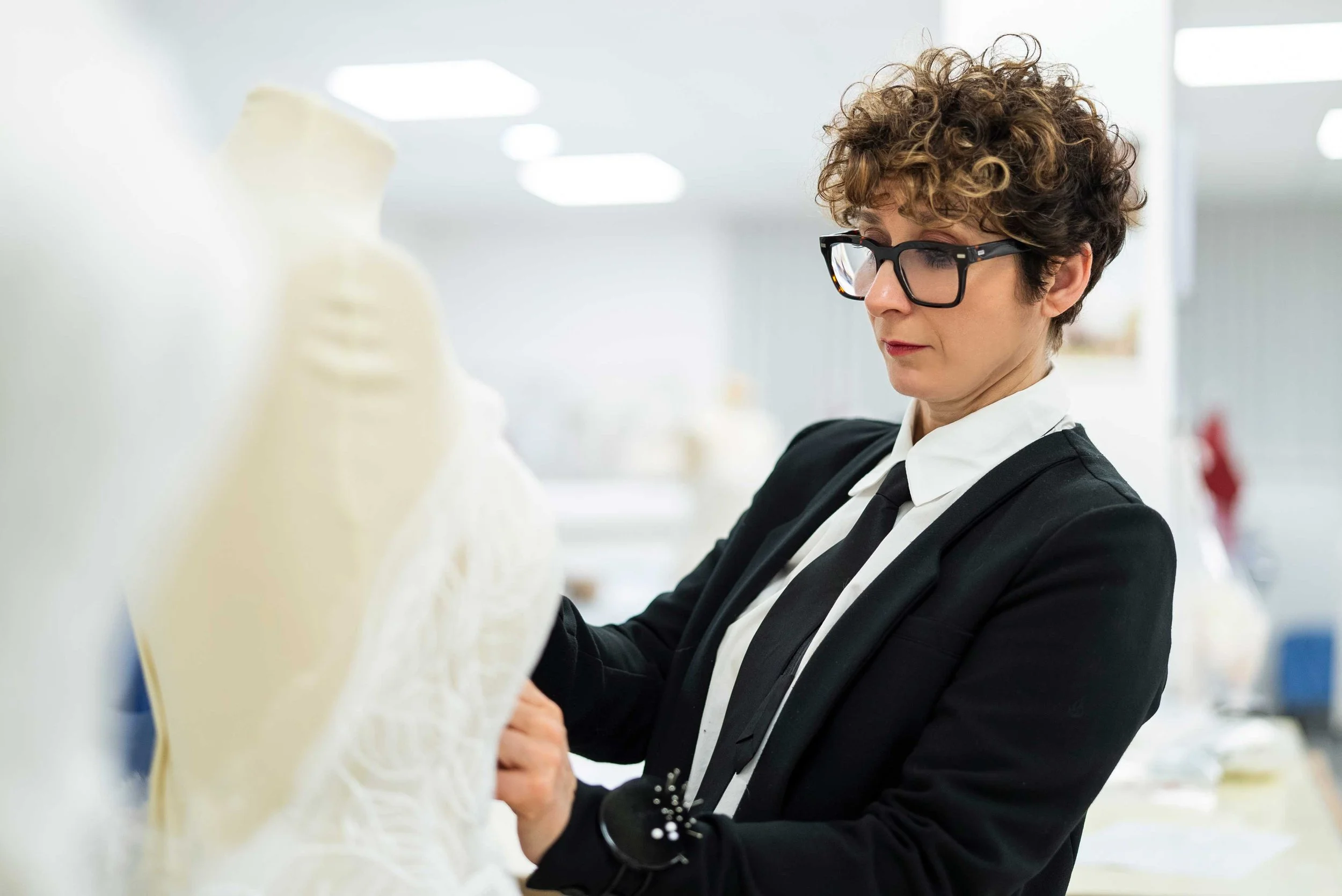 Valentini at New York Bridal Market. A woman with short curly hair, wearing glasses, a black blazer, white shirt, and black tie, examining a white bust sculpture in a well-lit workspace.