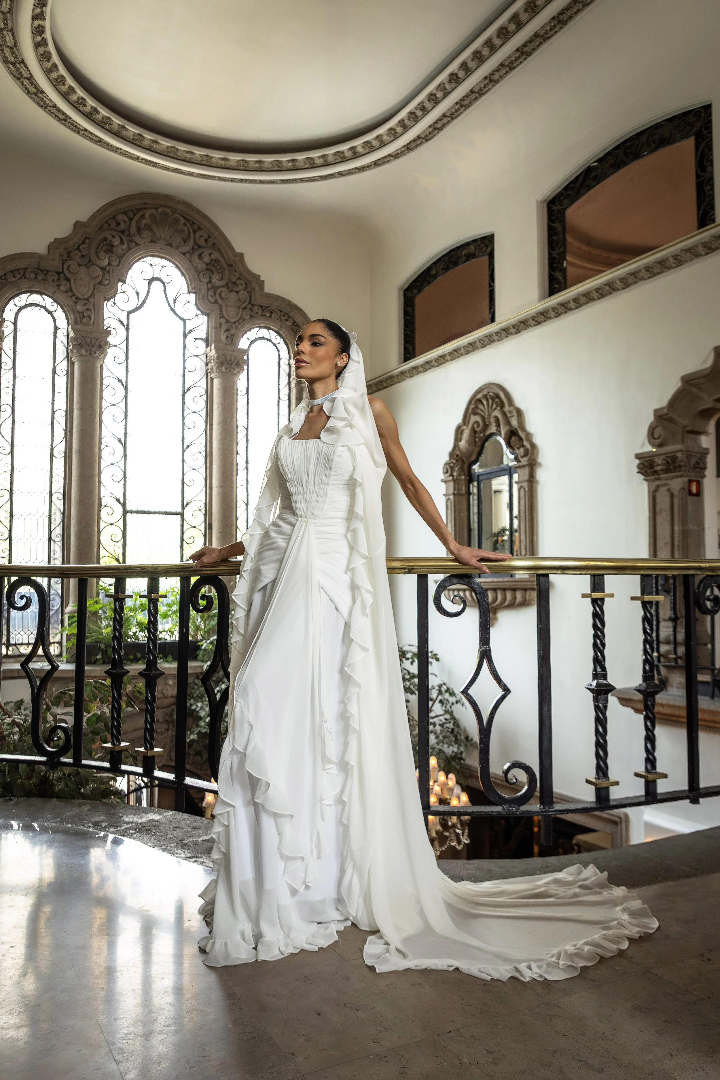 A woman in a white wedding dress with ruffles and a veil standing indoors near large arched stained glass windows, with candles and plants visible below the railing.