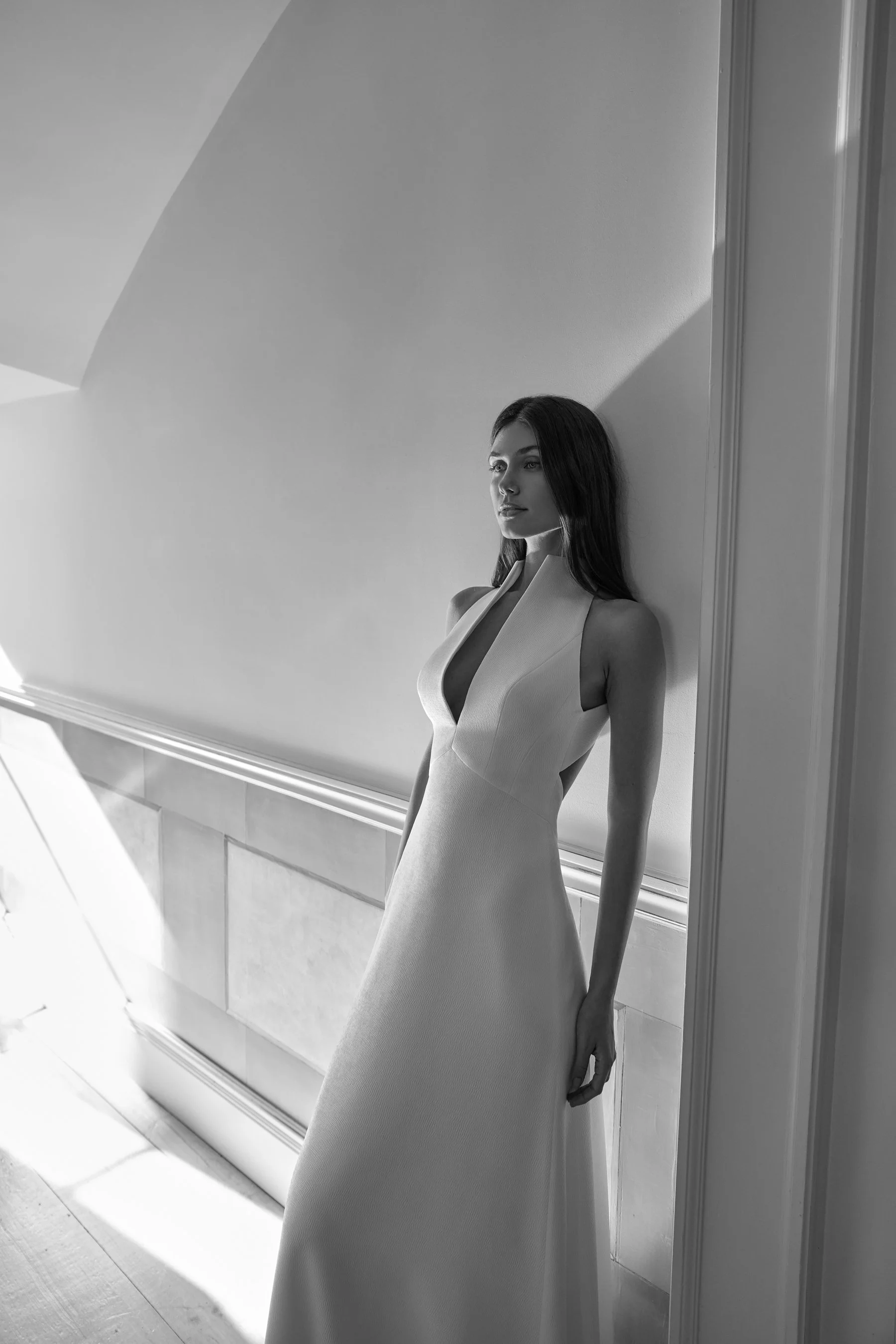 Black and white photo of a woman with long dark hair wearing a sleeveless, V-neck gown, standing beside a wall with a shadow cast at an angle. She is looking away from the camera, appearing contemplative.