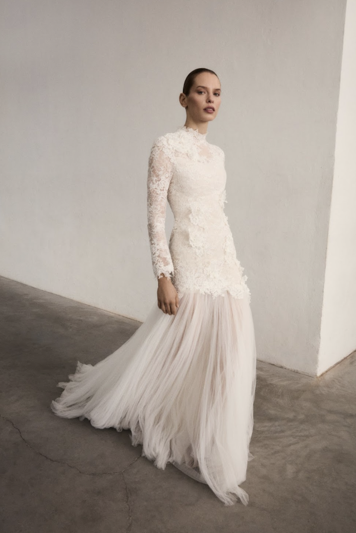 Marmar Halim at New York Bridal Market A woman in a long white lace wedding dress with a pleated tulle skirt stands against a plain, light-colored wall.