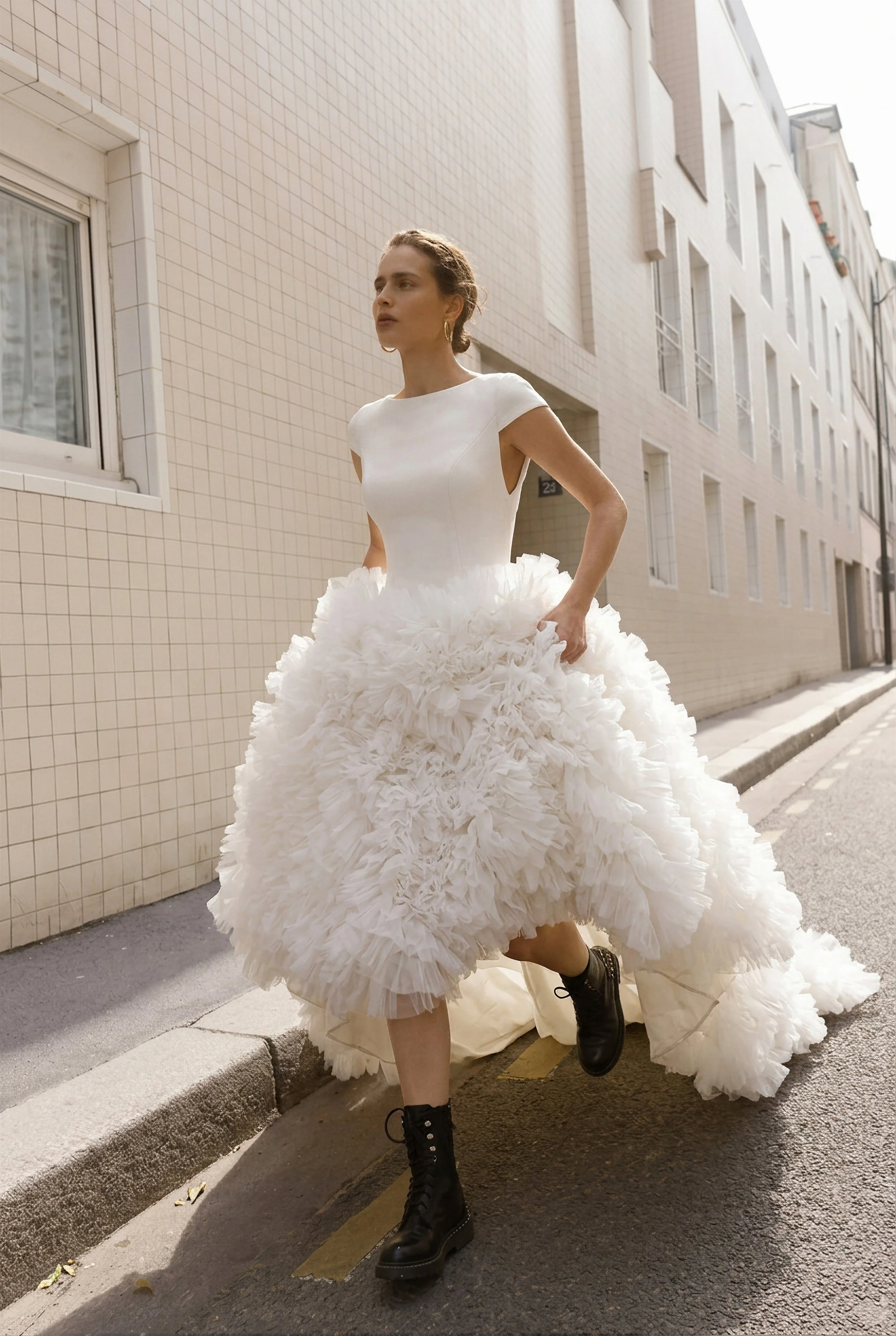 Gemy Maalouf New York Bridal Market A young woman wearing a white, strapless dress with a large, textured skirt, black combat boots, and gold hoop earrings, standing on a city street with beige tiled buildings in the background.