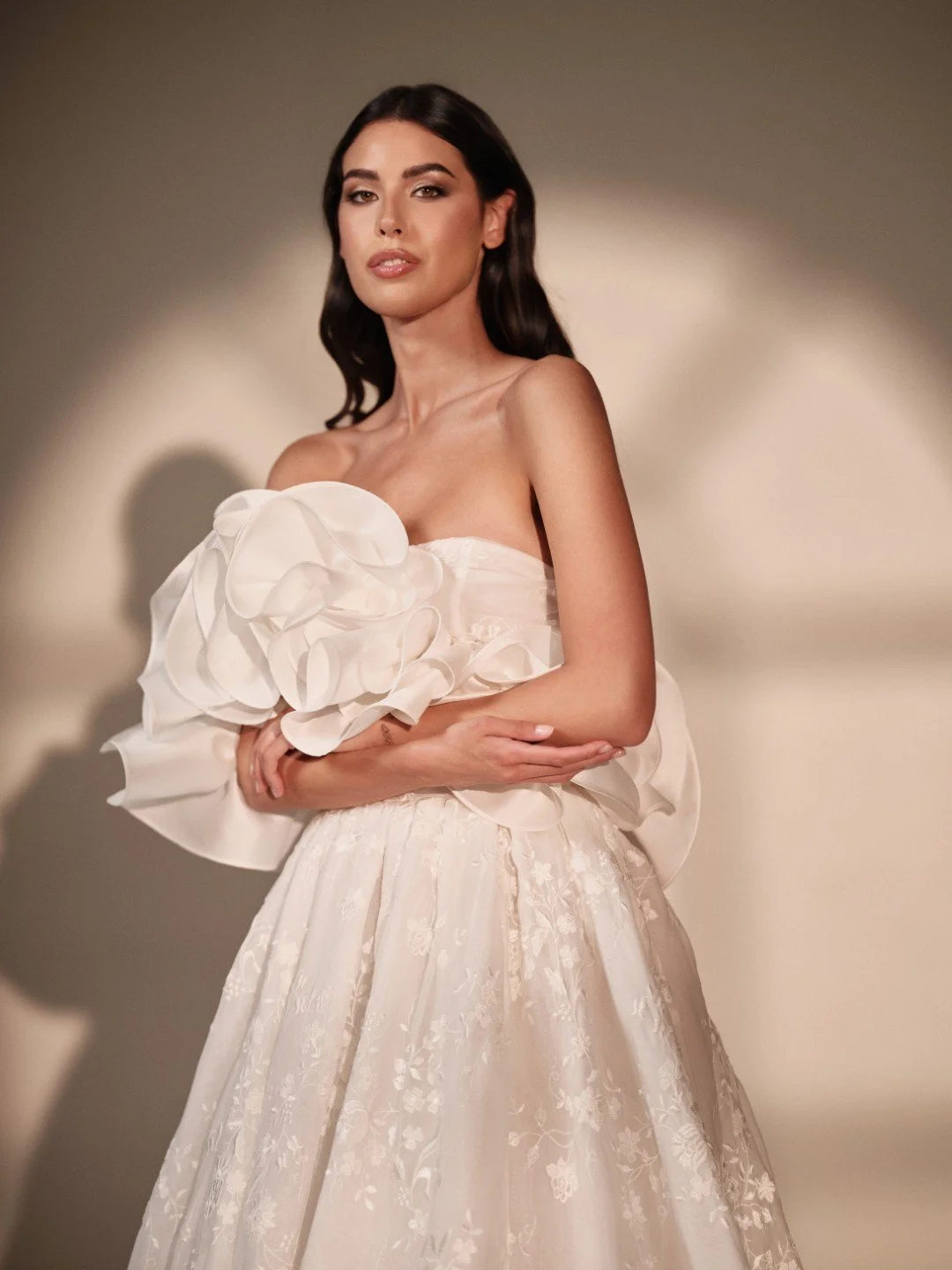 Maria Pia at New York Bridal Market.  A woman with dark hair in a strapless white dress with ruffled floral details on the bodice, holding a matching flower-embellished accessory, standing against a neutral background.