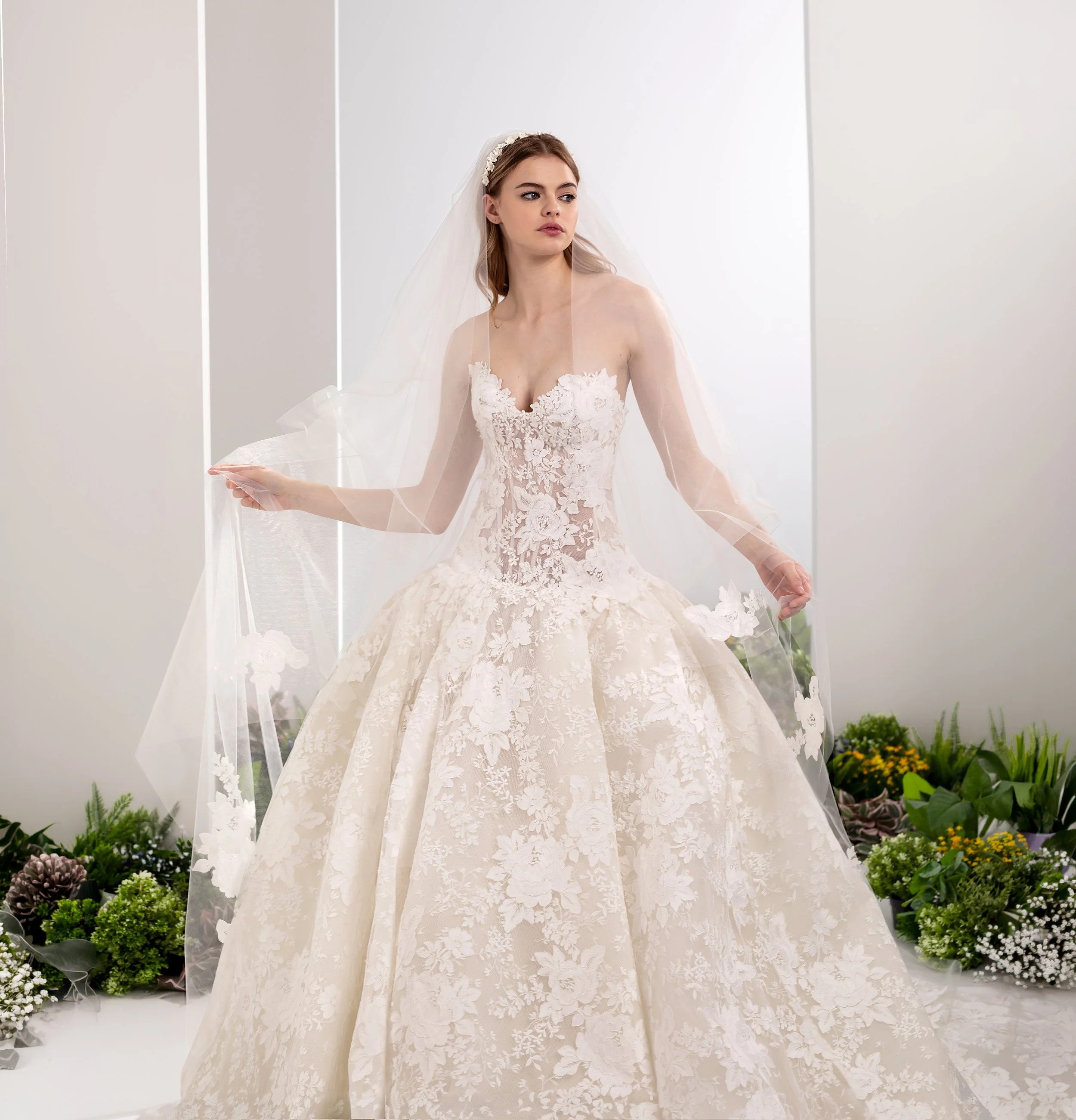 Valentini Spose at New York Bridal Market.  A bride in a white wedding dress with floral lace details and a sheer veil, standing in front of a white background with plants and flowers at her feet.