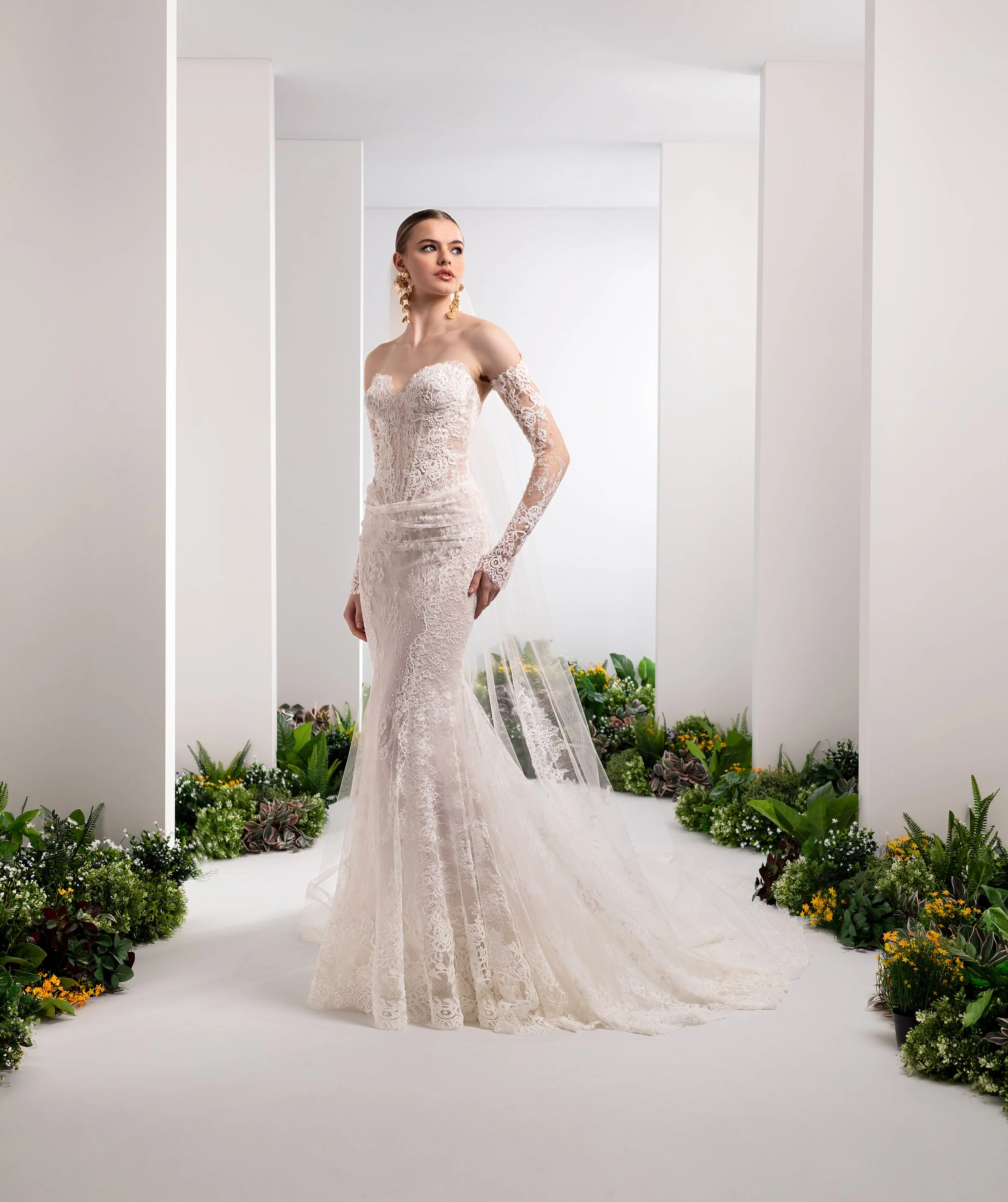Valentini Spose at New York Bridal Market.  A woman in a white lace wedding dress with long sleeves, standing in a minimalist, white venue decorated with green plants and yellow flowers.