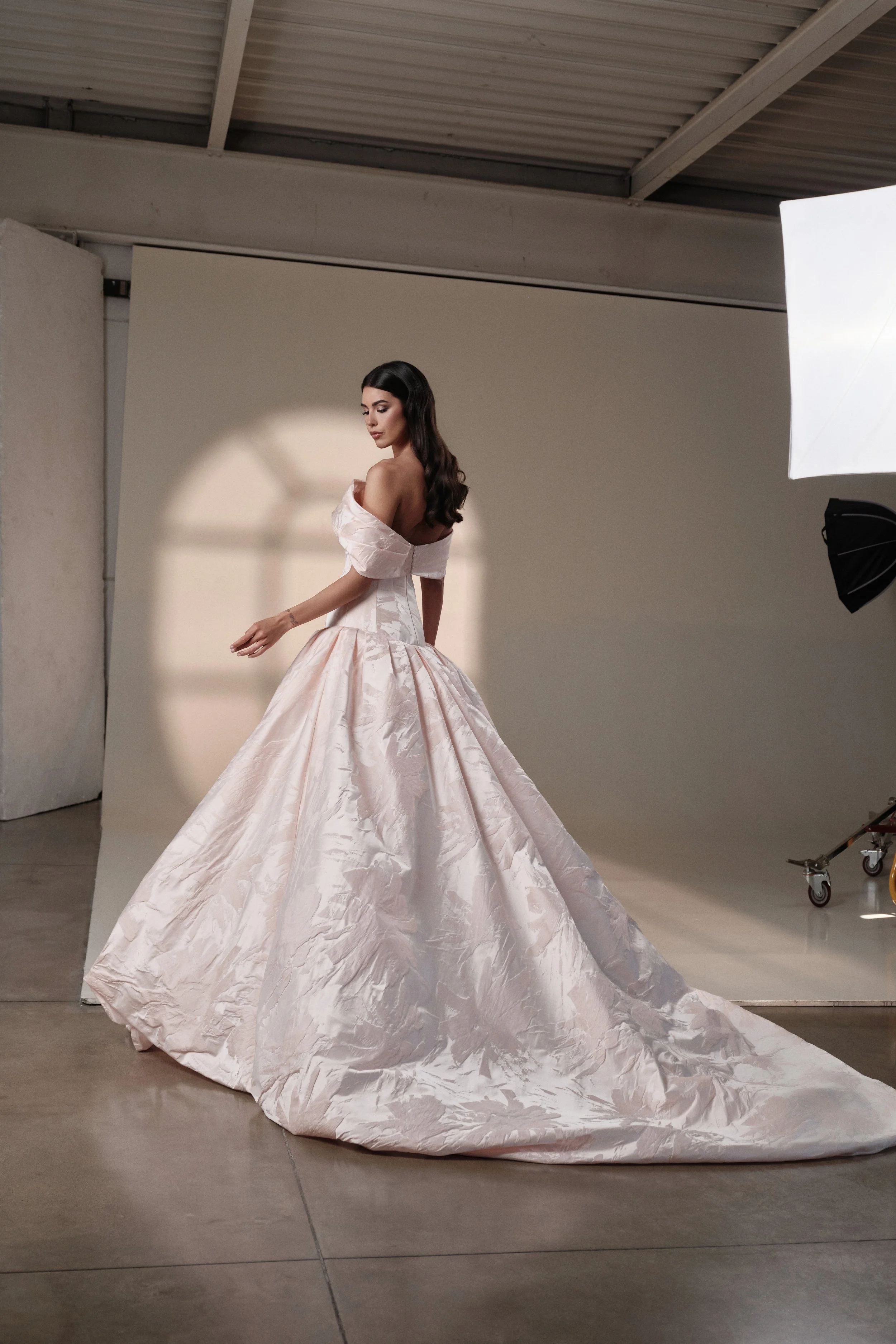 A woman in a luxurious off-shoulder, light-colored gown with a full skirt in a photography studio, standing against a beige background with studio lighting and equipment visible.