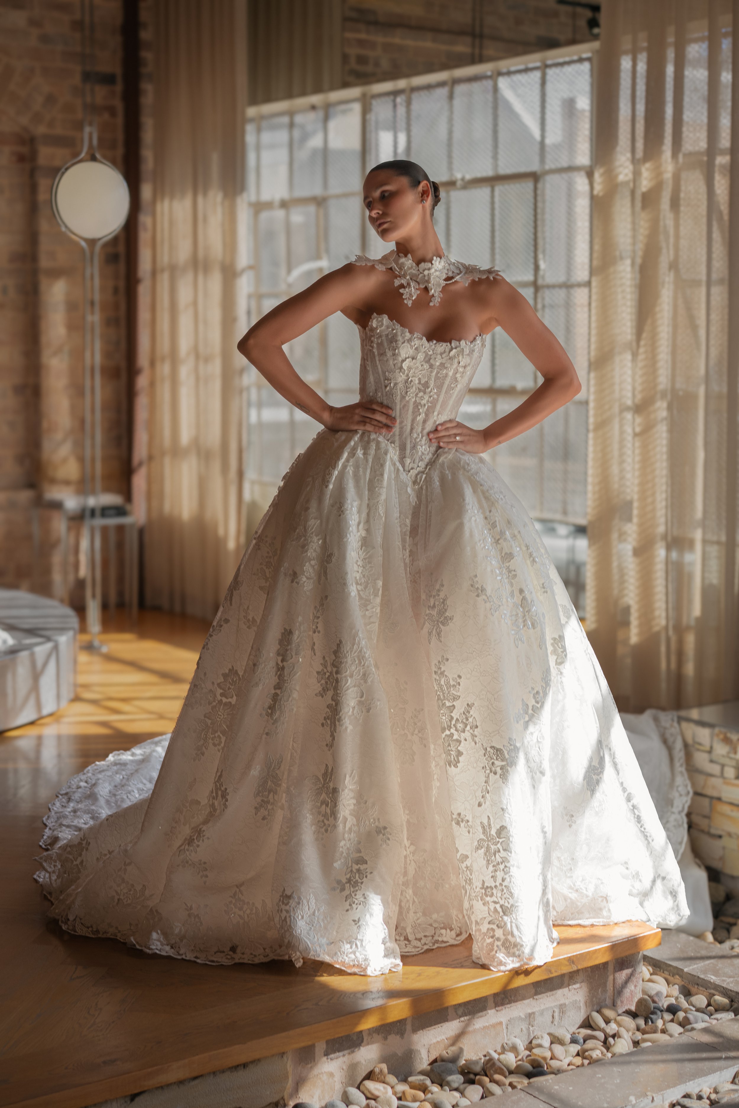 Blanche Bridal at New York Bridal Market  A woman wearing an elegant, lace wedding dress stands in a warmly lit industrial-style room with large windows and exposed brick walls.