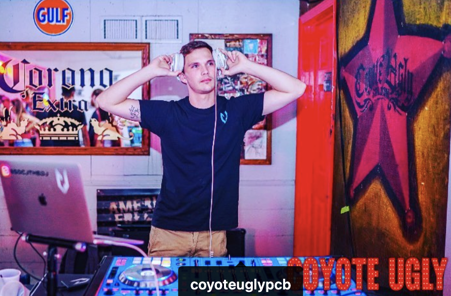 A young man wearing headphones stands behind DJ equipment in a bar or club setting, with neon and graffiti-style posters on the wall.