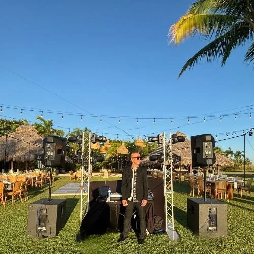 Man in black suit and sunglasses standing in front of DJ equipment at outdoor event under string lights, with thatched-roof structures and palm trees in the background.